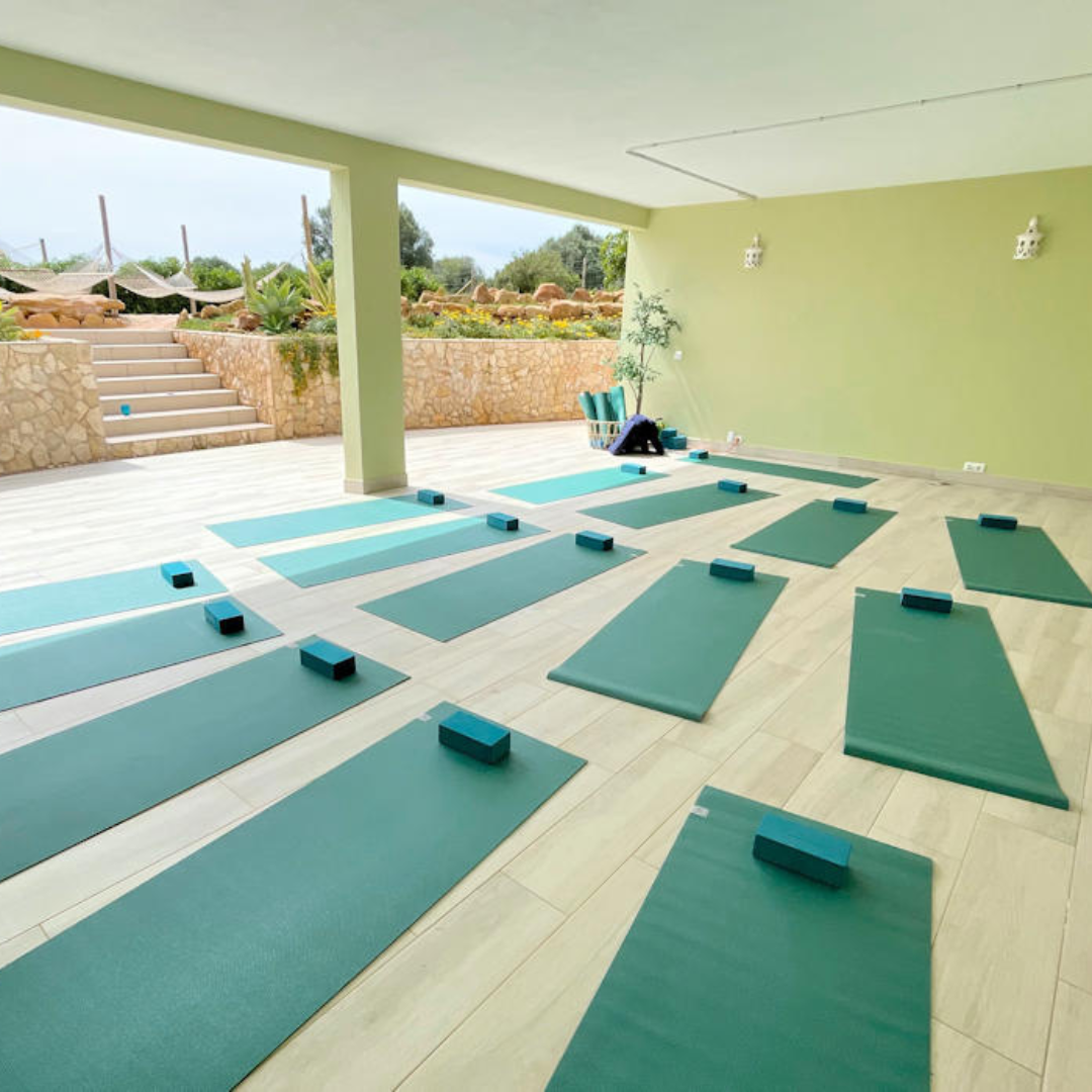 Outdoor yoga space with green mats and blocks on a tiled floor, surrounded by stone walls and greenery.