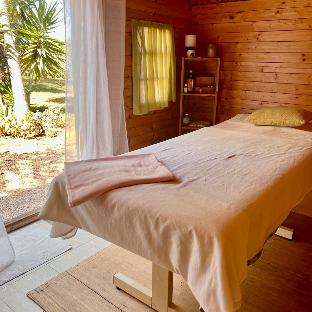 Cozy spa room with wooden walls, massage table, soft linens, and a view of a garden through sheer curtains. Shelves with wellness products are in the corner.