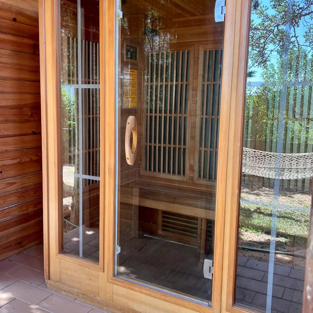 Wooden infrared sauna with glass door, located outside on a tiled floor, surrounded by trees.