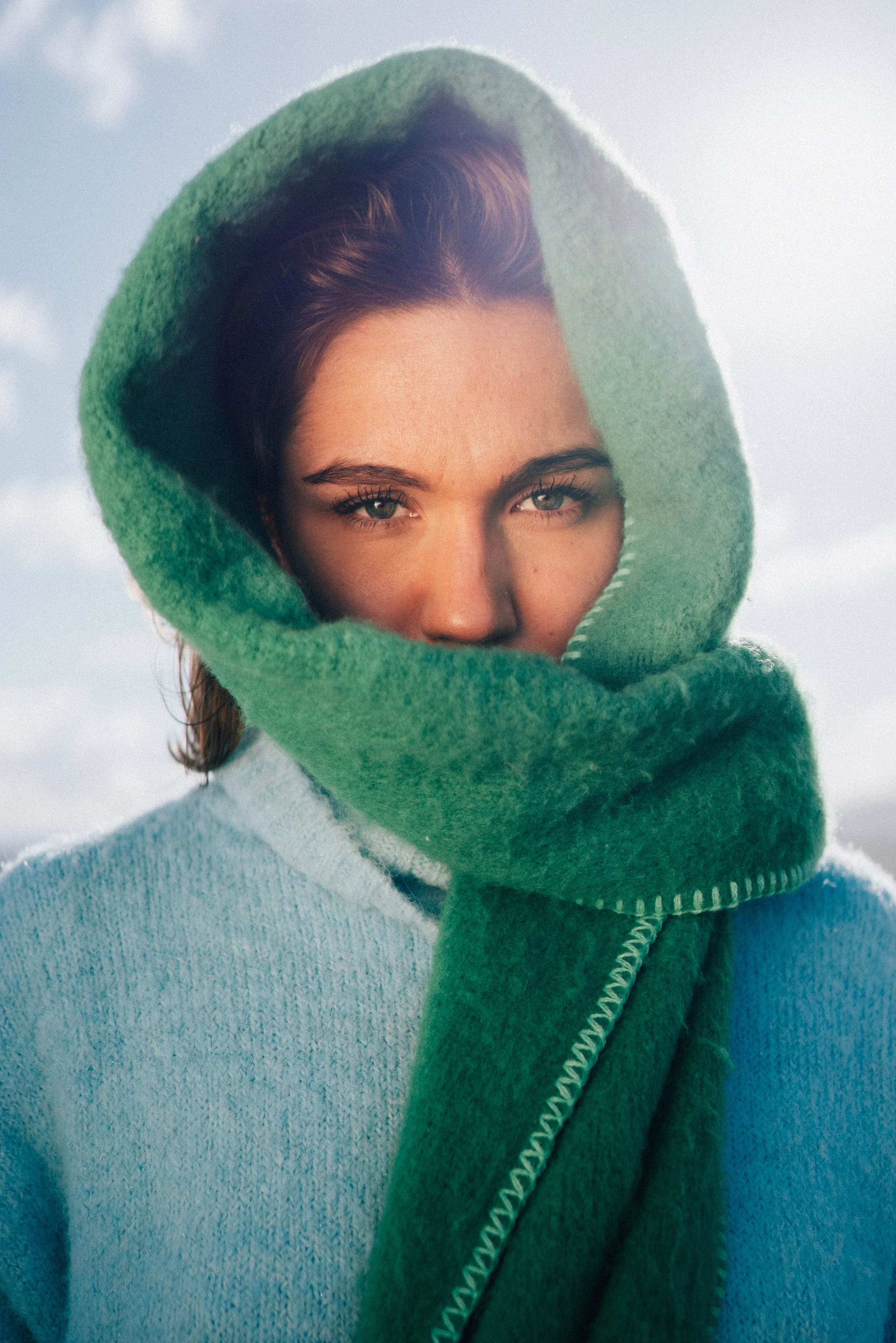 A woman wearing a blue sweater and a large green scarf wrapped around her head and neck, outdoors with cloudy sky background.