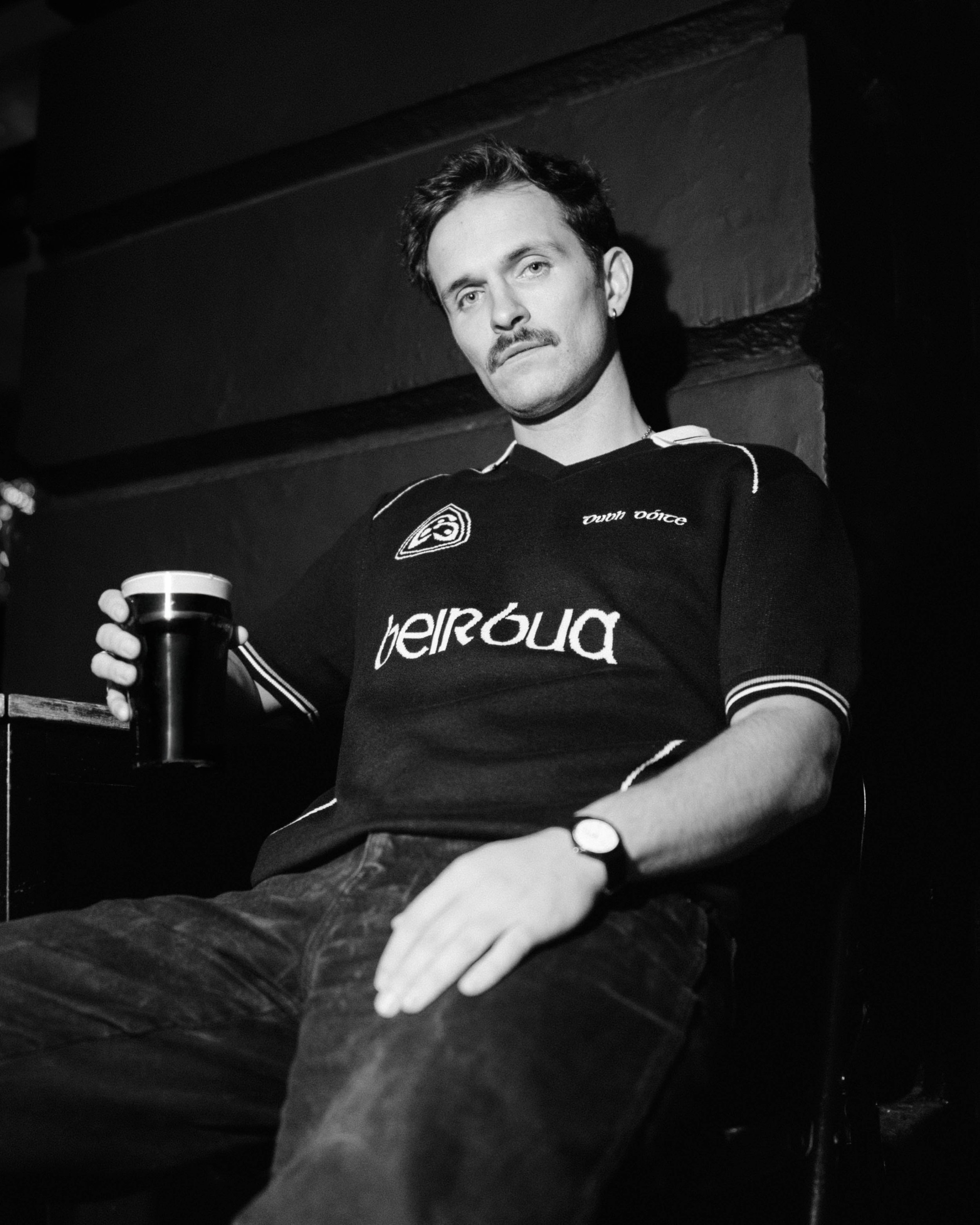 A man with a mustache sitting in a dimly lit room, holding a cup of dark beverage, wearing a sports jersey with a watch on his left wrist, seated against a brick wall.