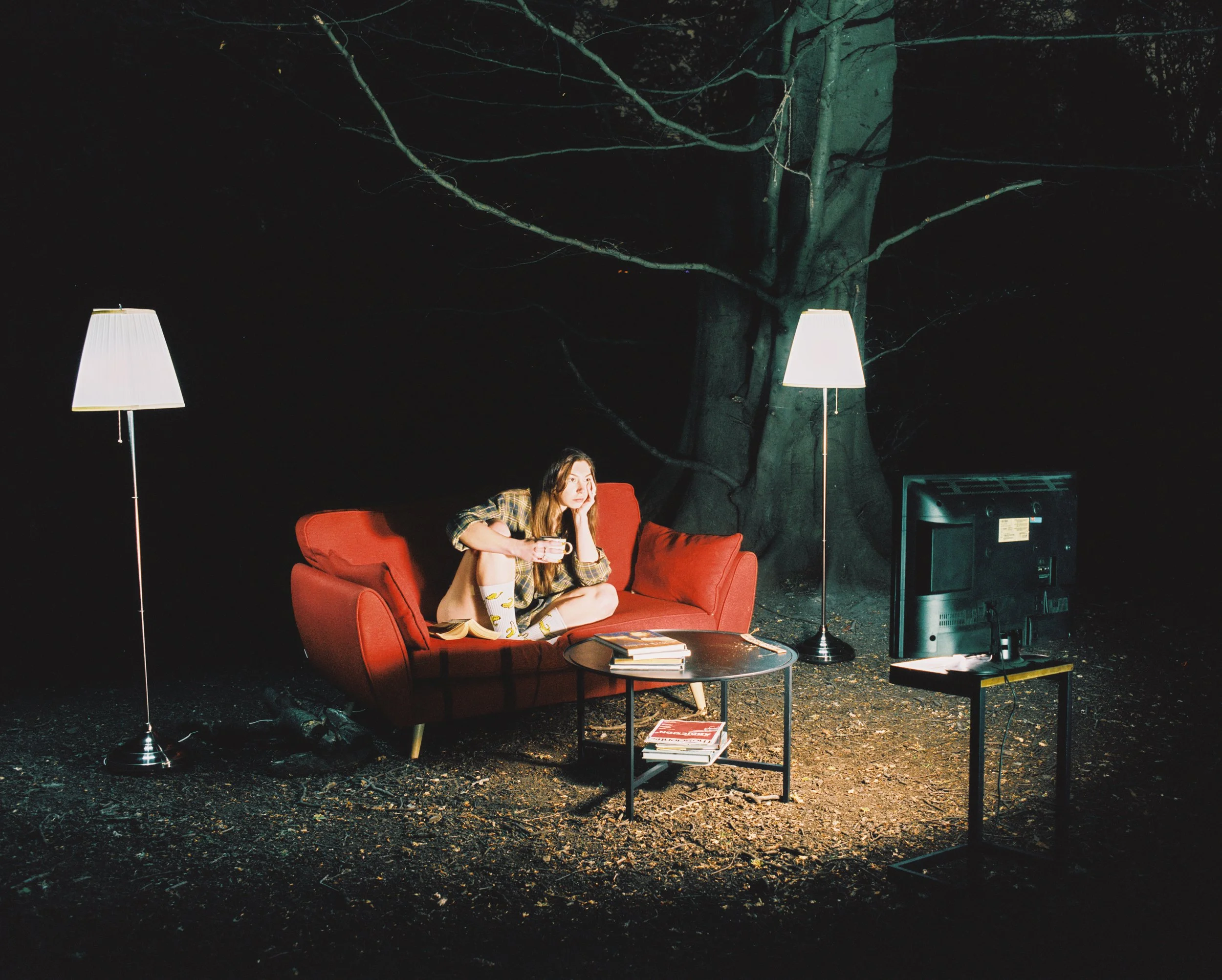 A woman sitting on a red sofa outdoors at night, watching television with two tall floor lamps on either side and a small table with magazines in front, amidst a dark wooded area.