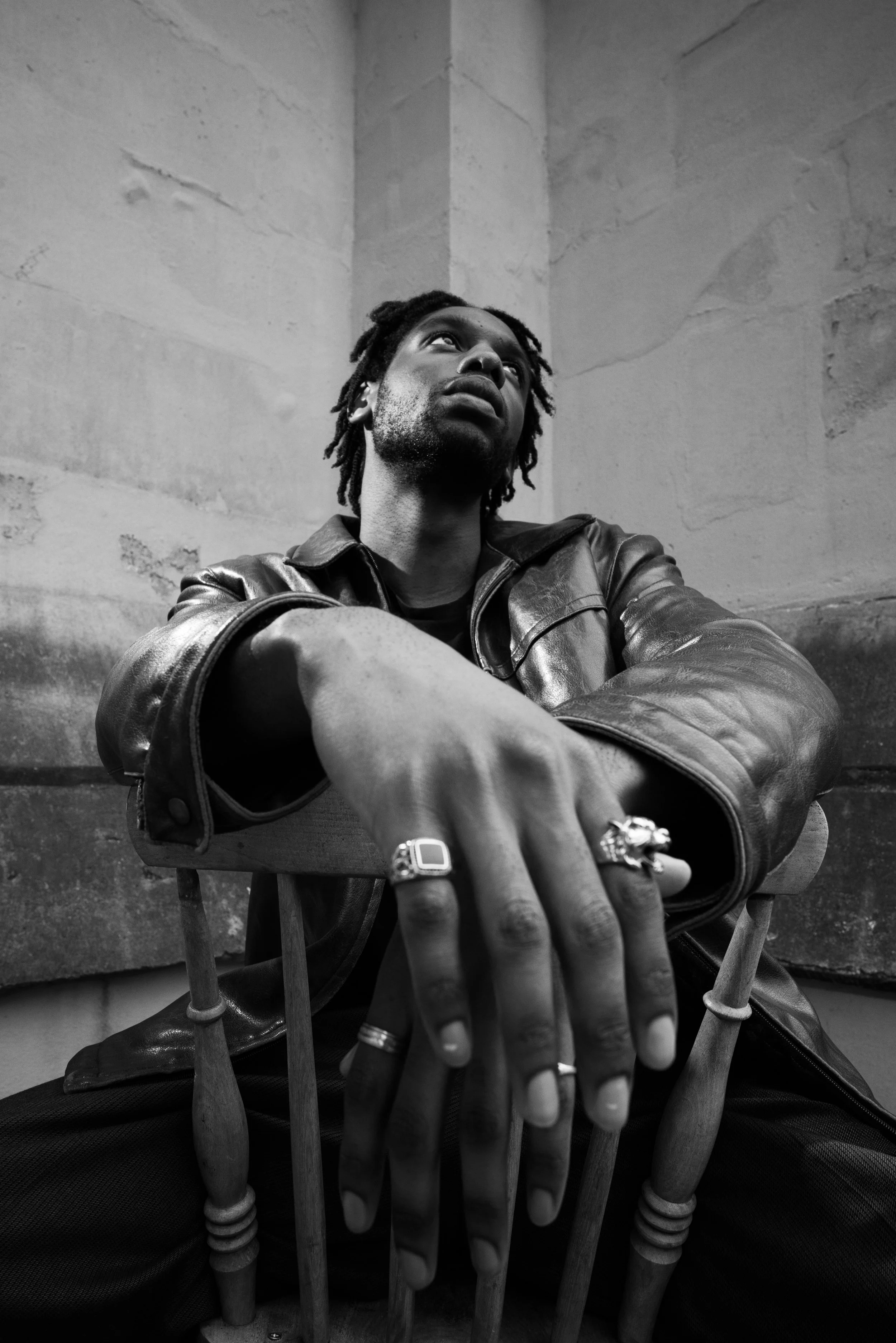 Black and white photo of a man sitting in a chair, wearing a leather jacket, with rings on his fingers, looking up with a thoughtful expression.
