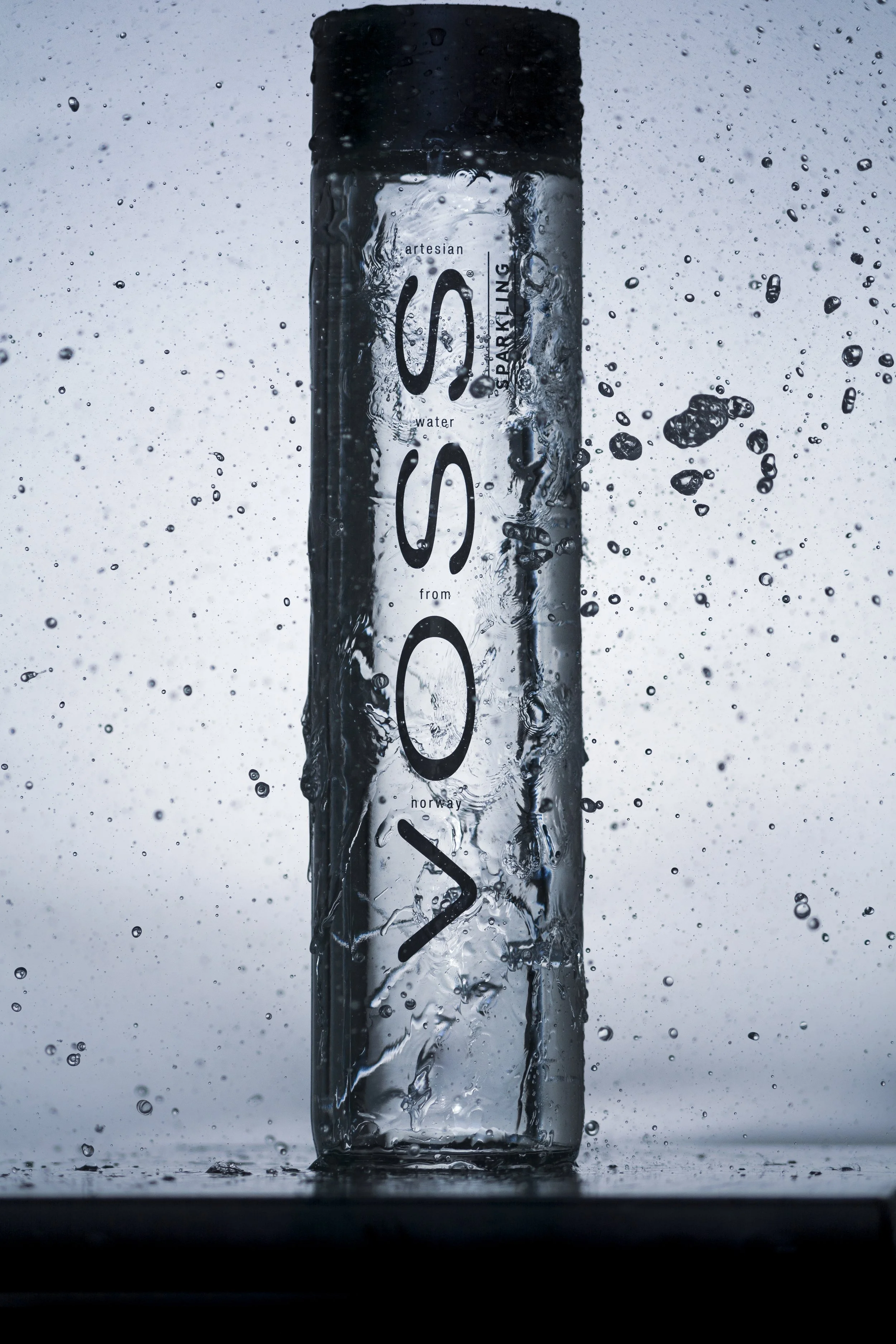 A clear plastic water bottle with black cap, labeled 'SYRO' and text indicating it is 'artesian water from Horway', surrounded by water droplets and splash.