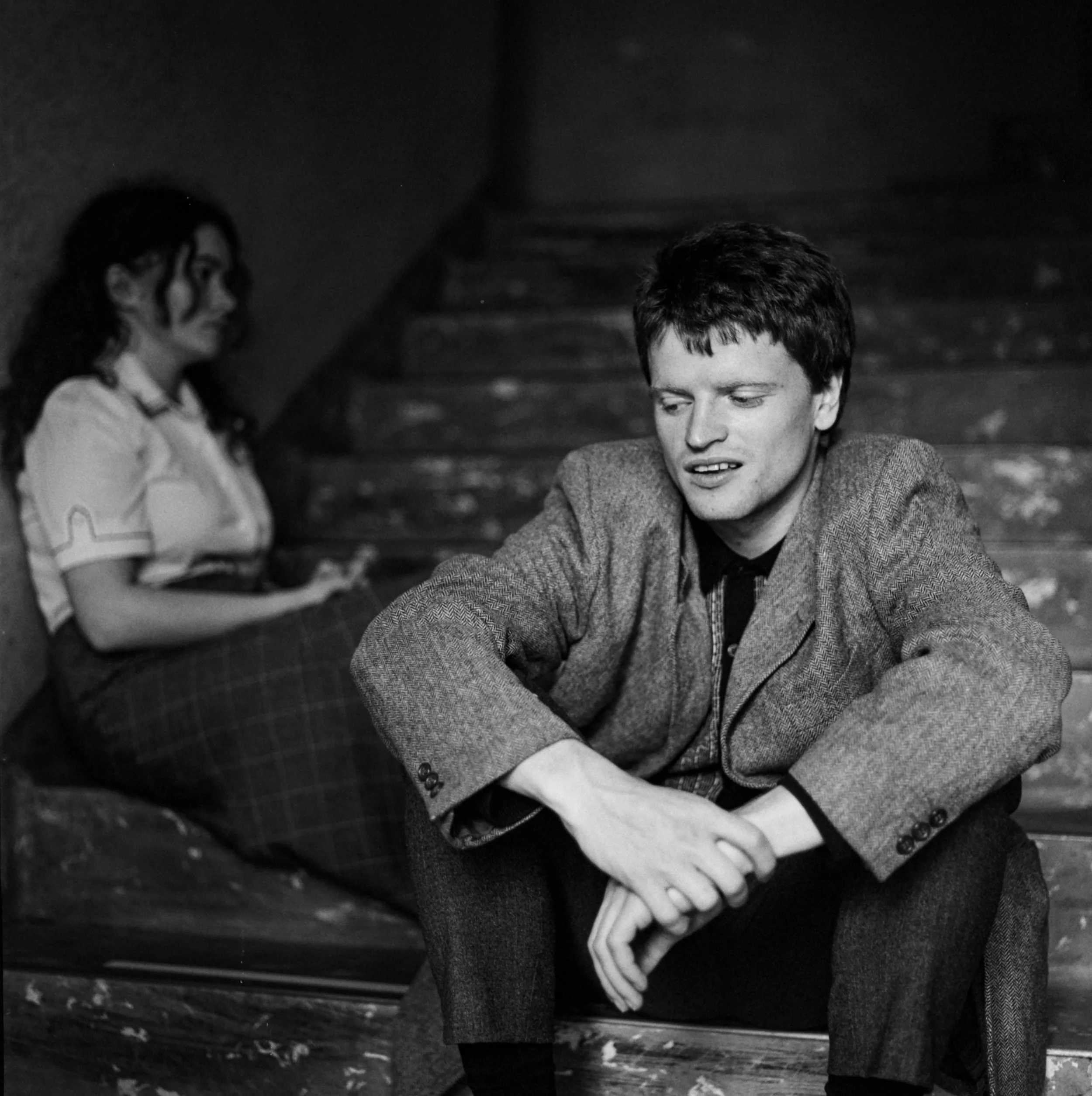 Black and white photo of a young man sitting on a step, looking distressed, with a young woman sitting on the stairs behind him, seemingly upset.