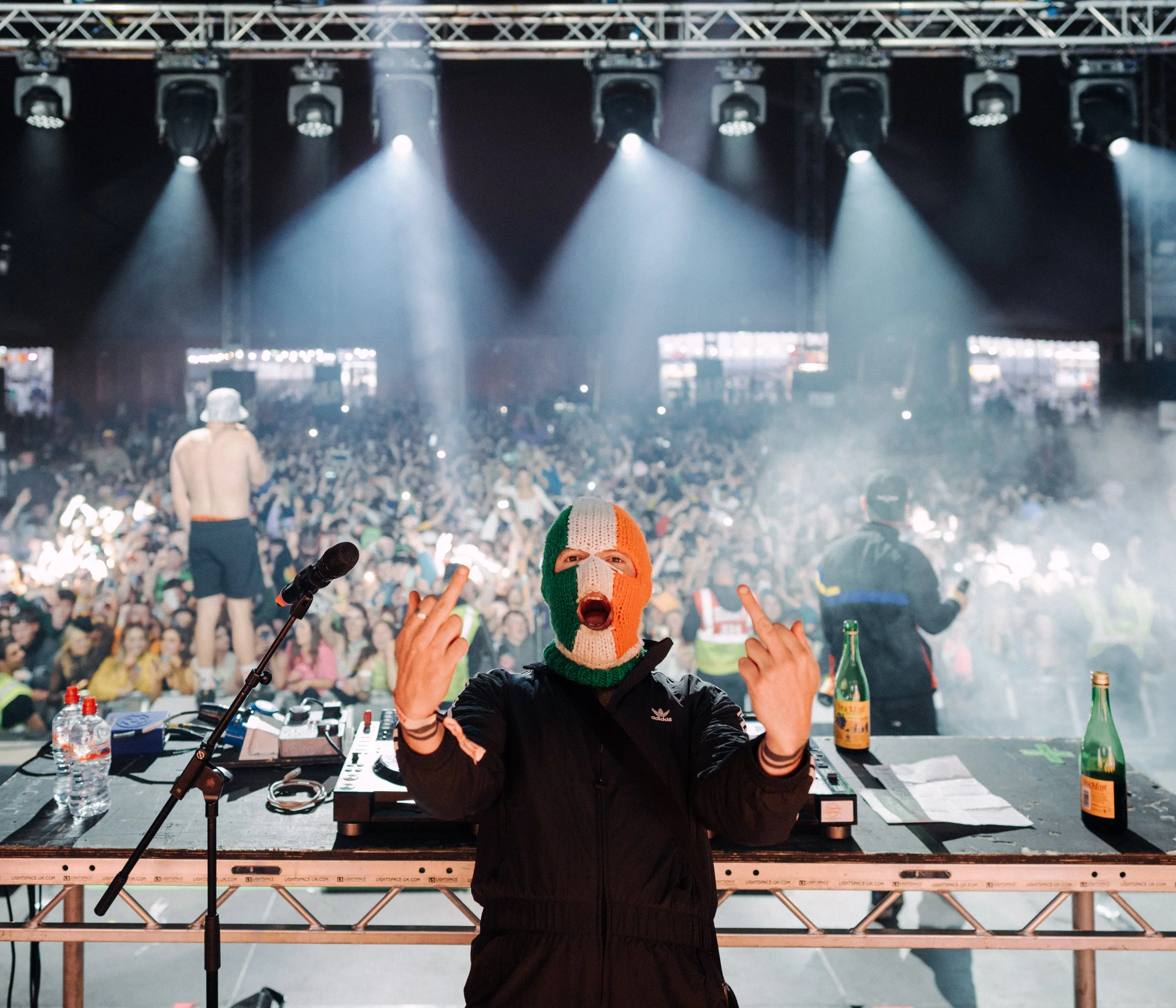 A DJ wearing an Italian flag ski mask and black jacket standing behind a DJ booth, making offensive hand gestures at a crowded concert or festival.