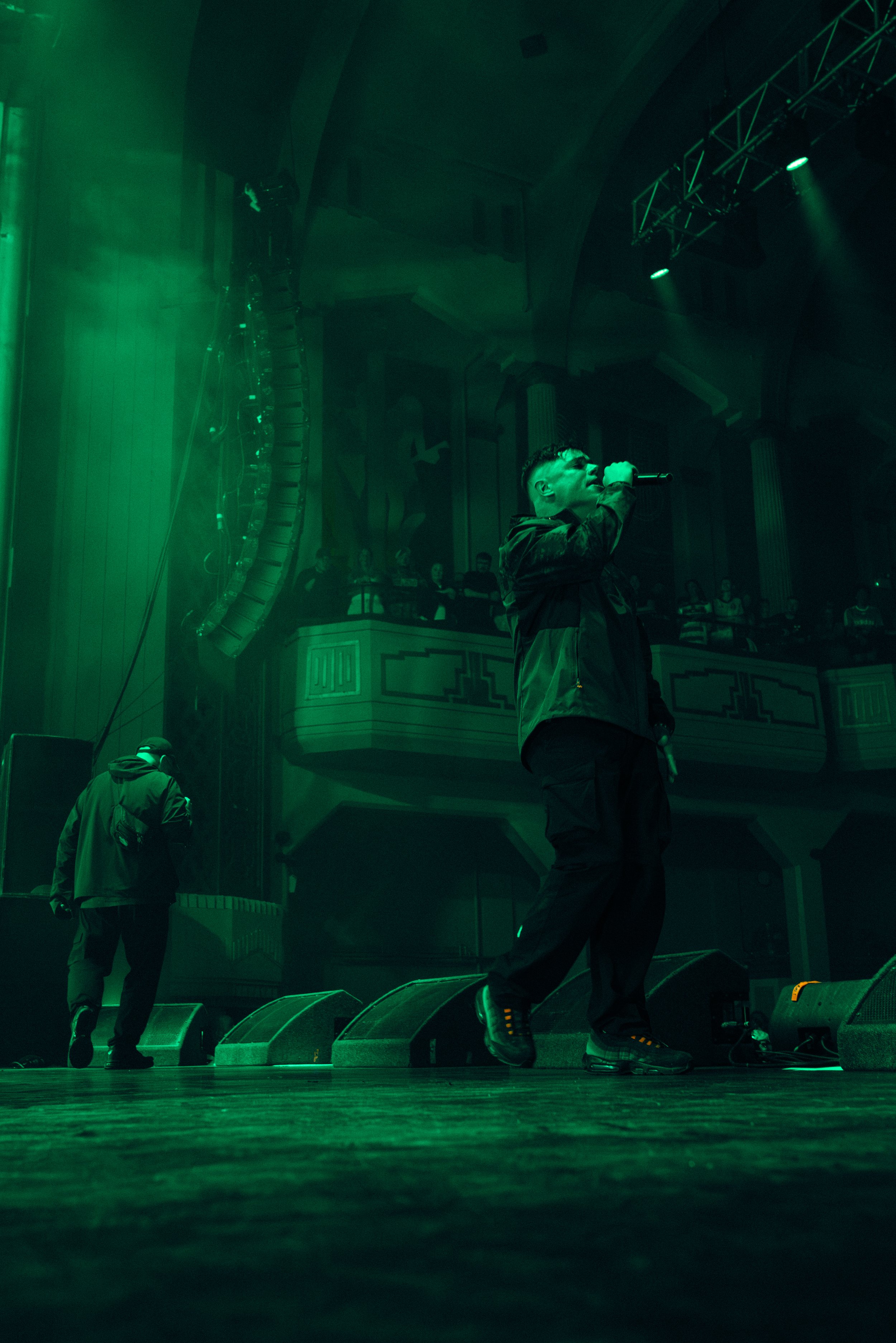 Performer singing into a microphone on stage in a dark concert hall with green lighting and audience in the background.