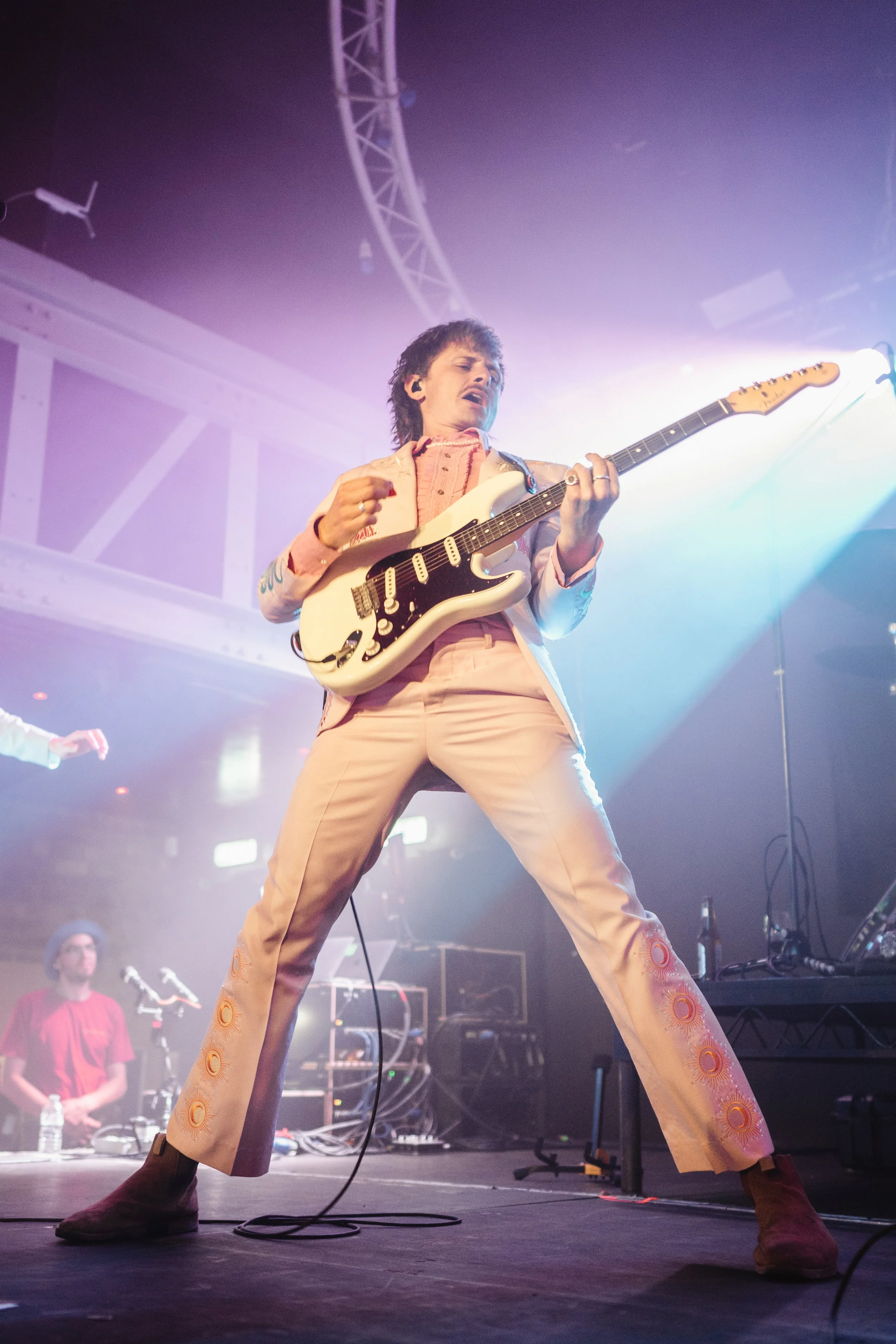 A male guitarist performing passionately on stage with colorful lighting, wearing a light pink suit and brown shoes, with a band member in the background.
