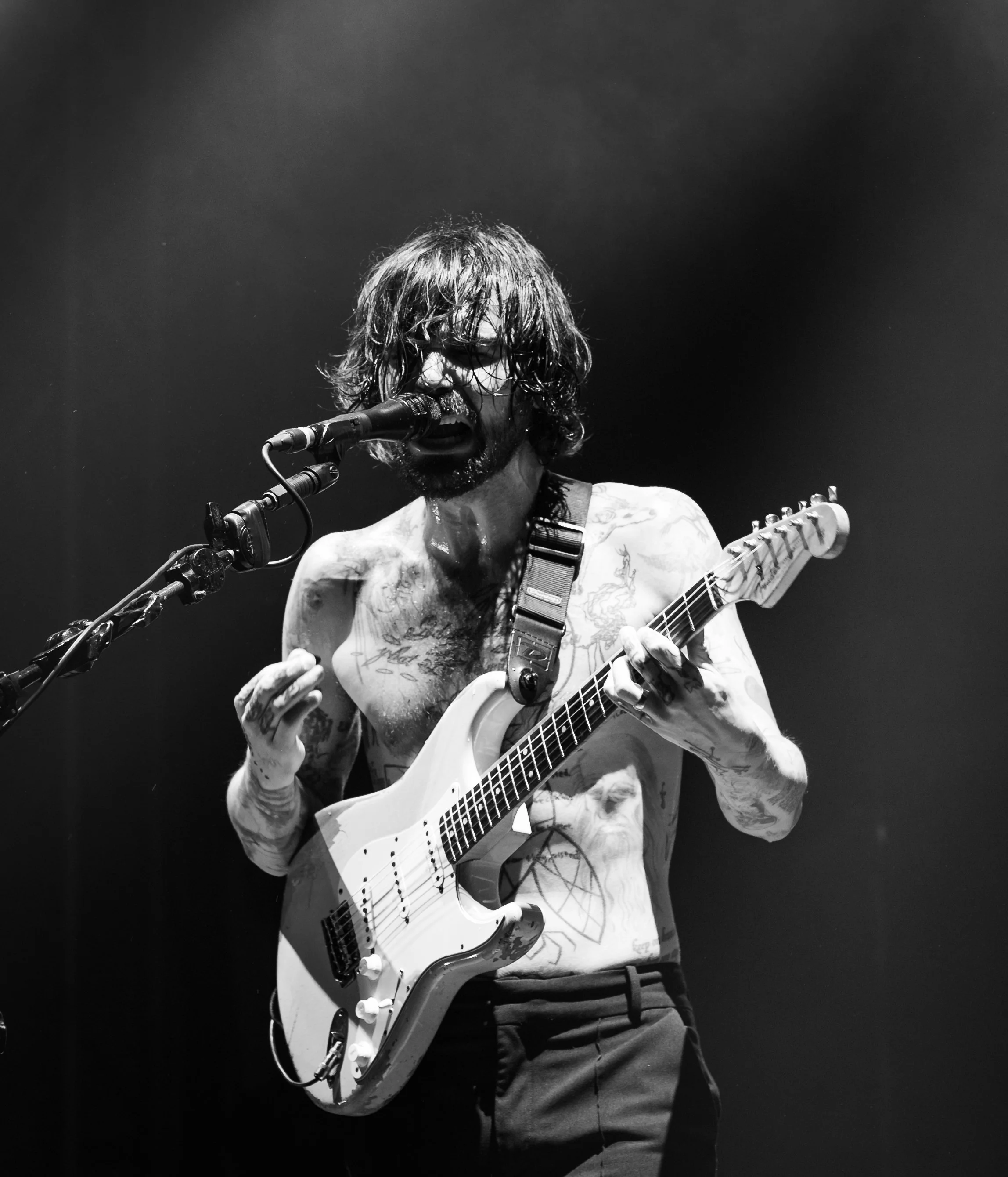 A shirtless man with tattoos, long hair, and glasses passionately playing an electric guitar and singing into a microphone on stage in black and white.