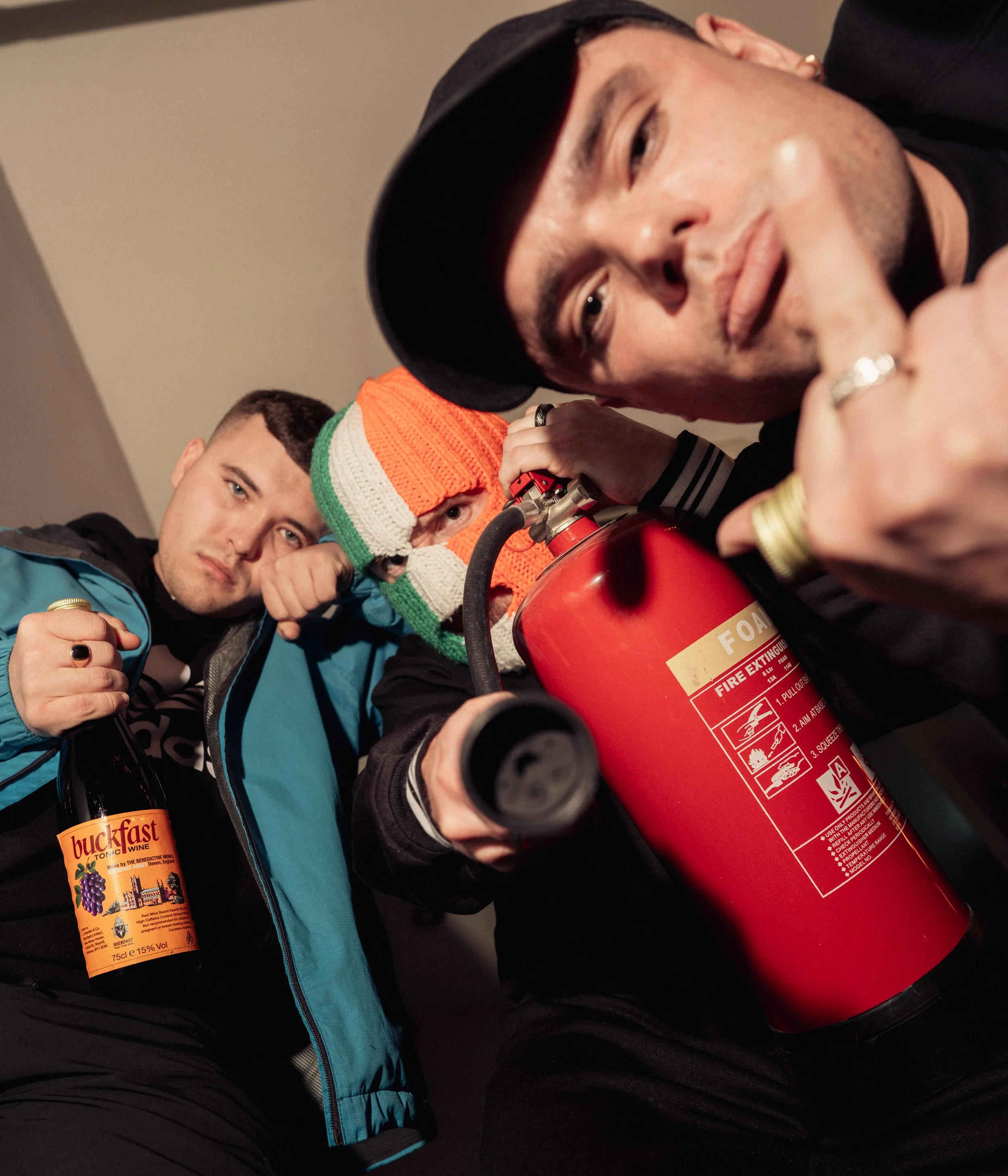 Three young men posing indoors. One is holding a bottle of Buckfast wine, another is holding a fire extinguisher, and the third man is touching his lips with his finger.