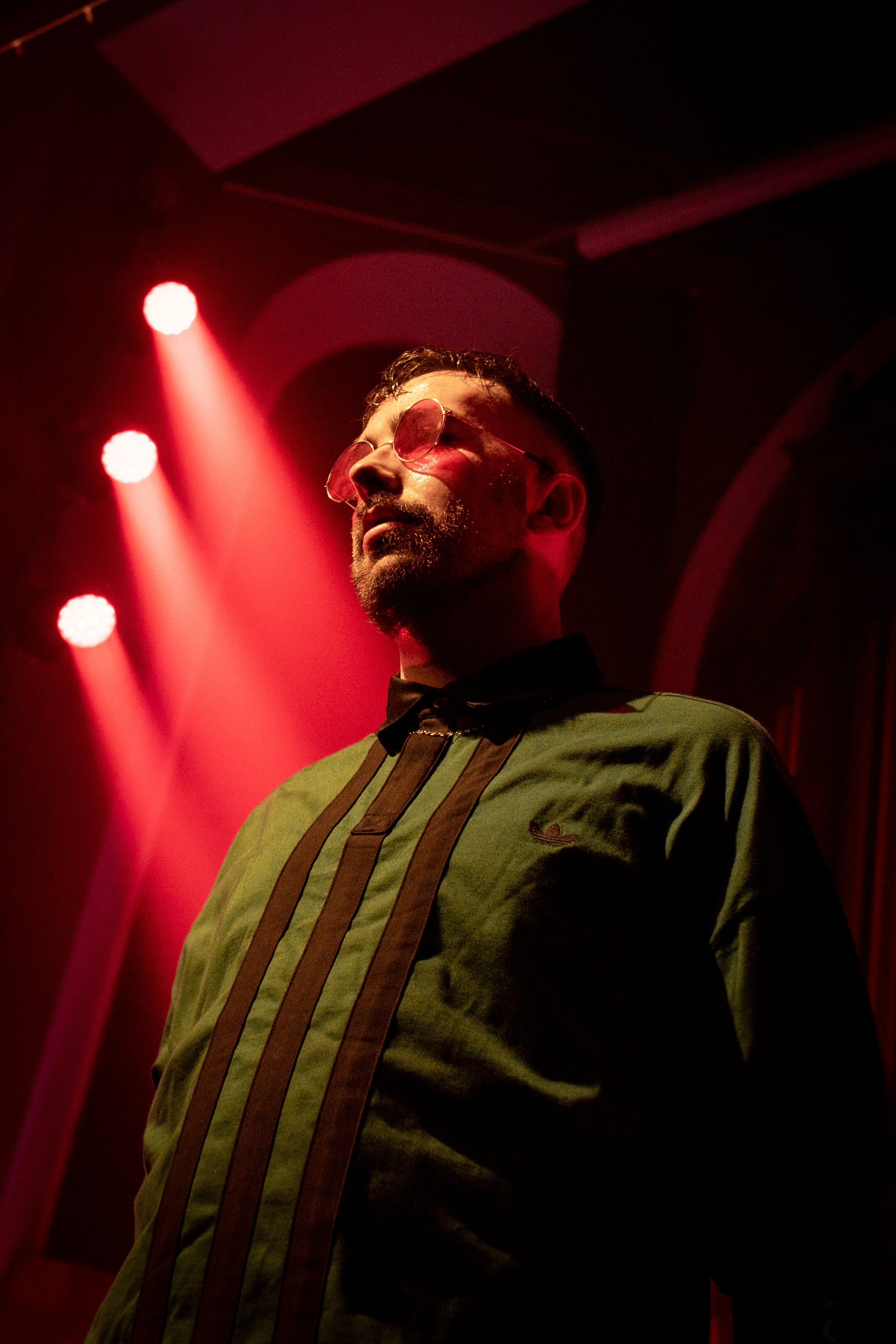 A man wearing round sunglasses and a green shirt with vertical black and brown stripes, standing on stage illuminated by red lights.