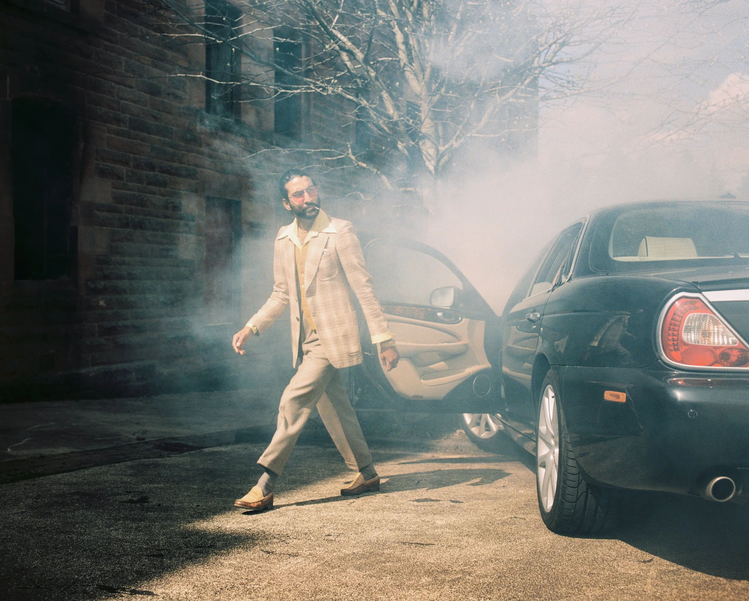 A man in vintage attire walking away from a black car with a door open, amid smoke or fog, on a city street with a brick building and tree in the background.