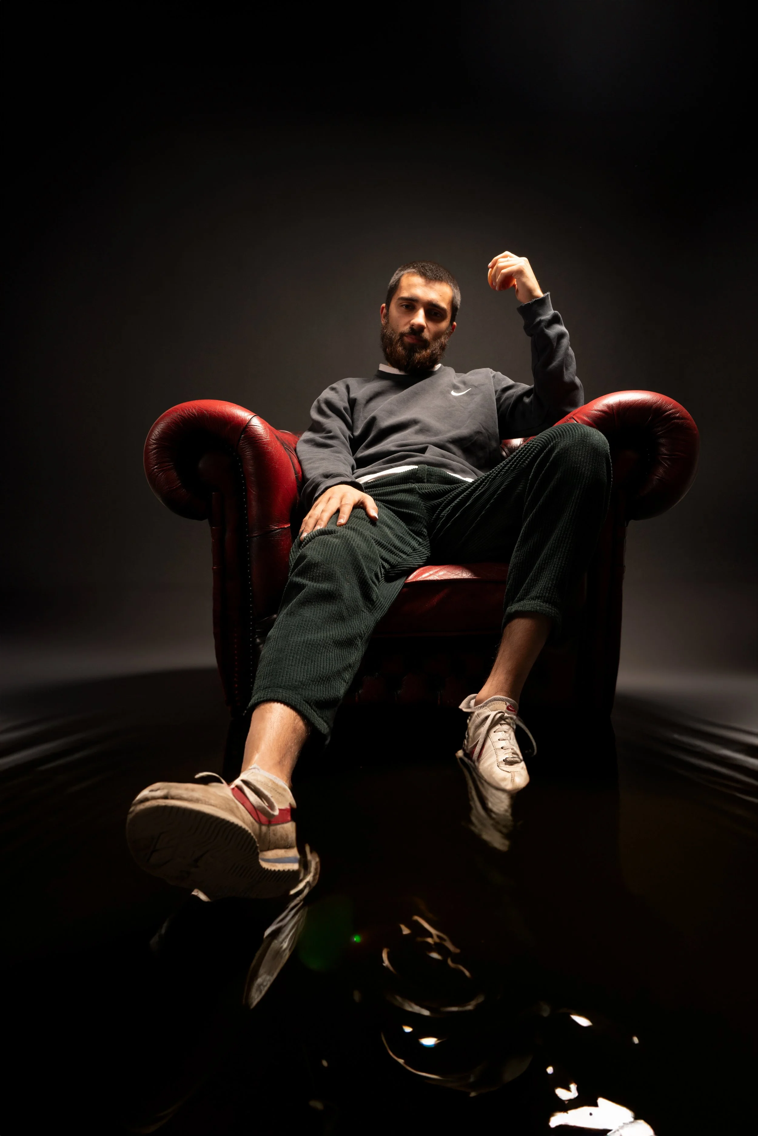 Man sitting in a red leather armchair against a dark background, wearing casual clothes, with one arm raised and a confident expression.