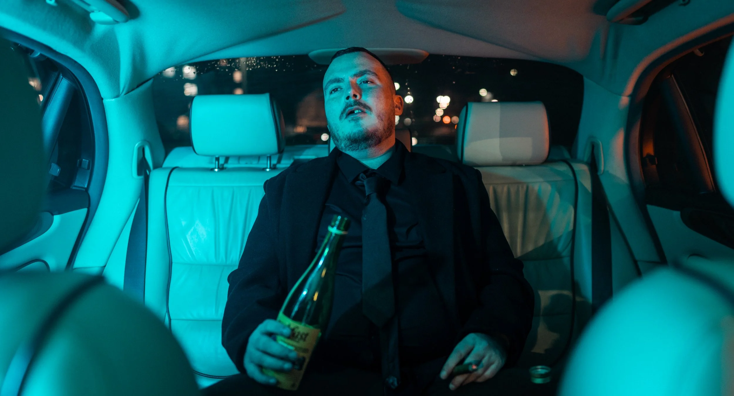 A man in a black suit and black tie sits in the backseat of a car at night, holding a bottle of beer in one hand and a cigarette in the other. The car's interior is lit with a teal hue, and city lights are visible through the rear window.