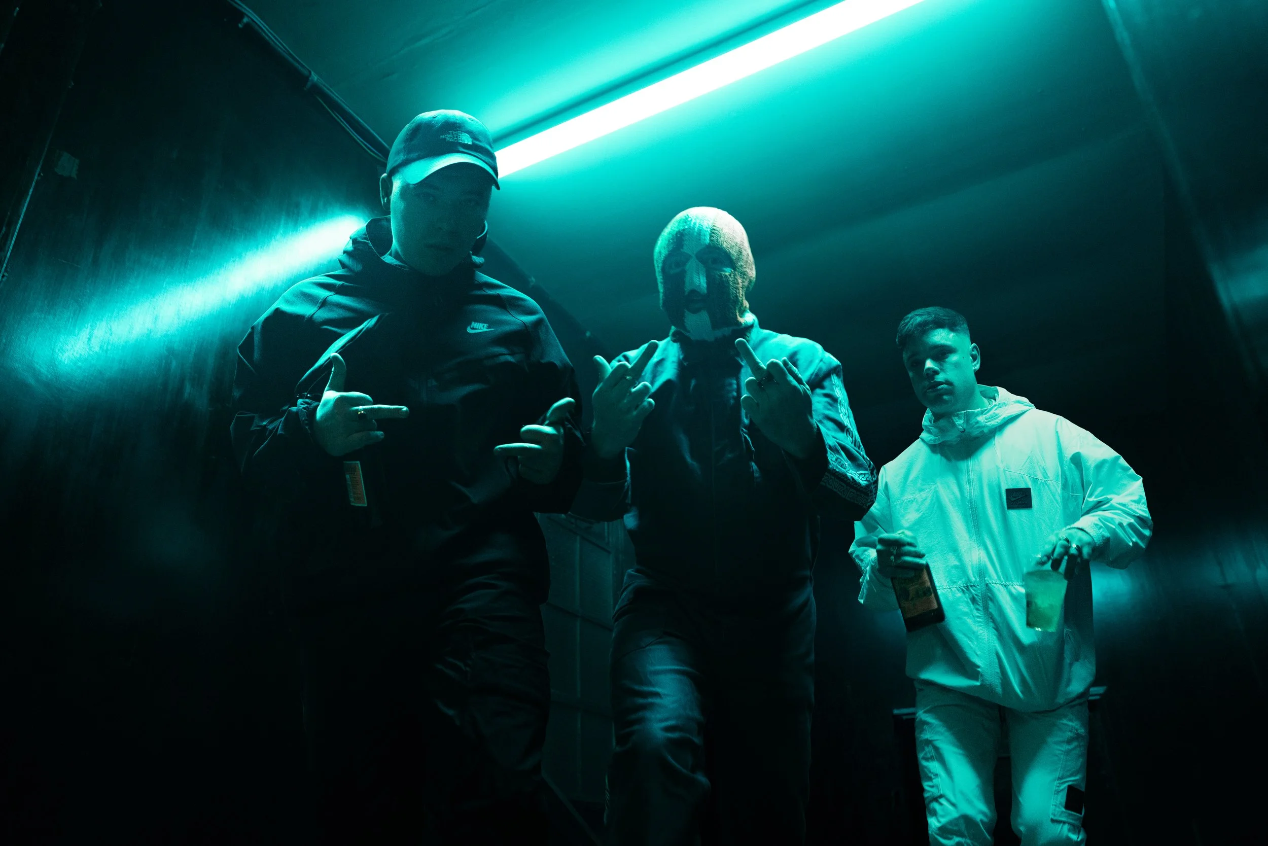 Three young men standing in a dark corridor with neon blue lighting. One wears a black jacket and cap, another has a skeleton mask and jacket, and the third is dressed in a white tracksuit.