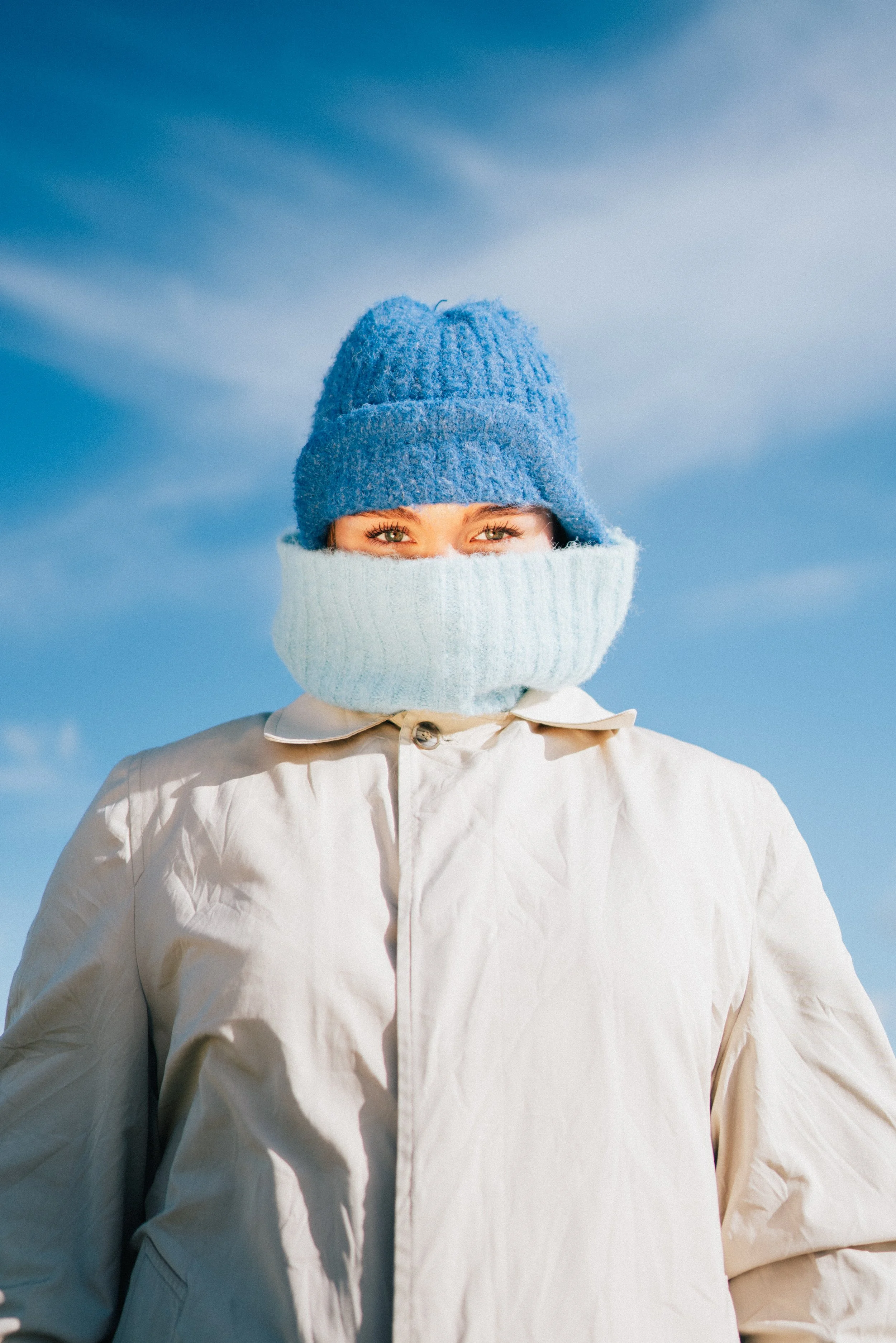 A woman bundled up in winter clothing, wearing a blue knit hat and a white scarf covering her mouth and nose, standing outdoors with a blue sky and wispy clouds in the background.