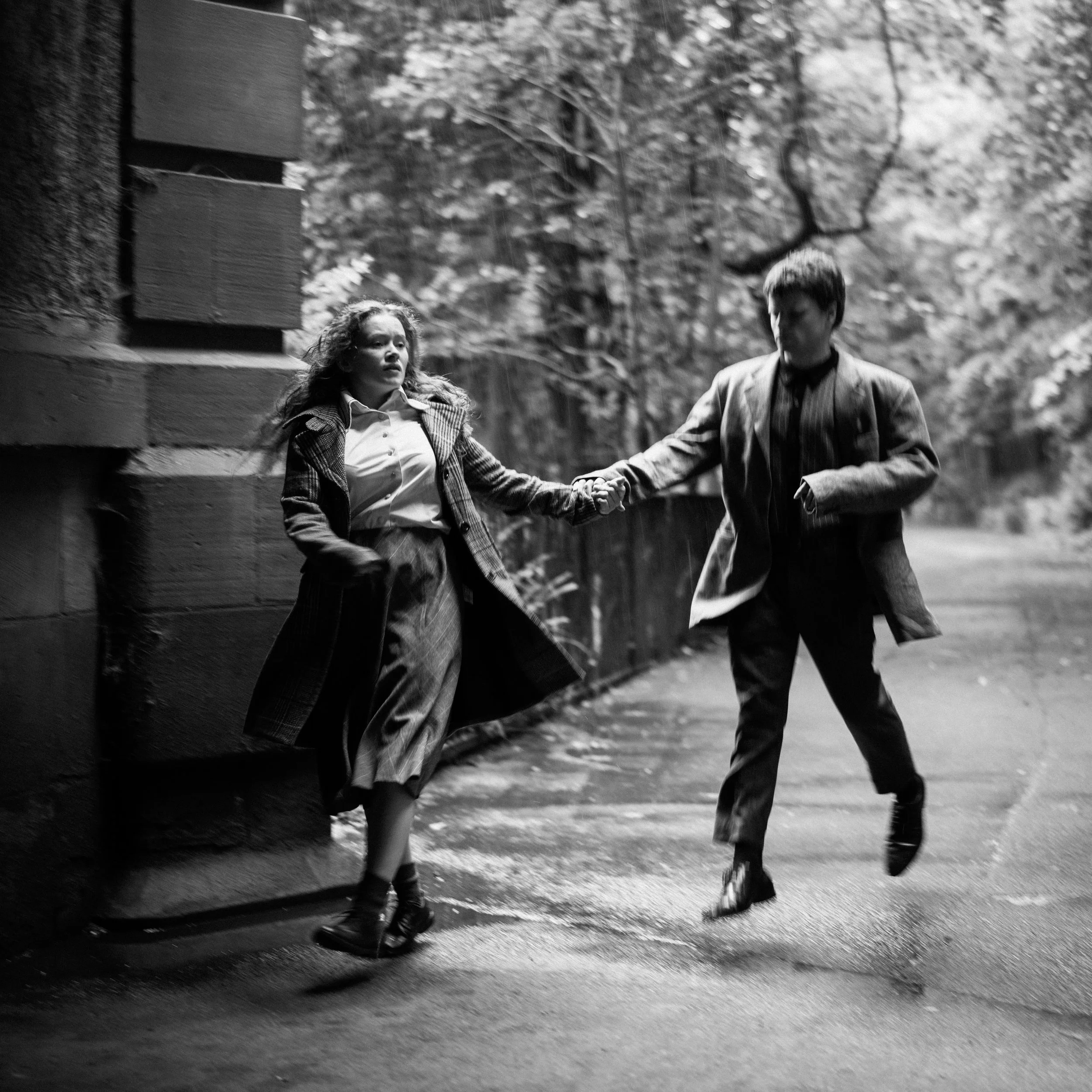 A black and white photo of a man and woman holding hands and walking together outdoors, with trees and a fence in the background.