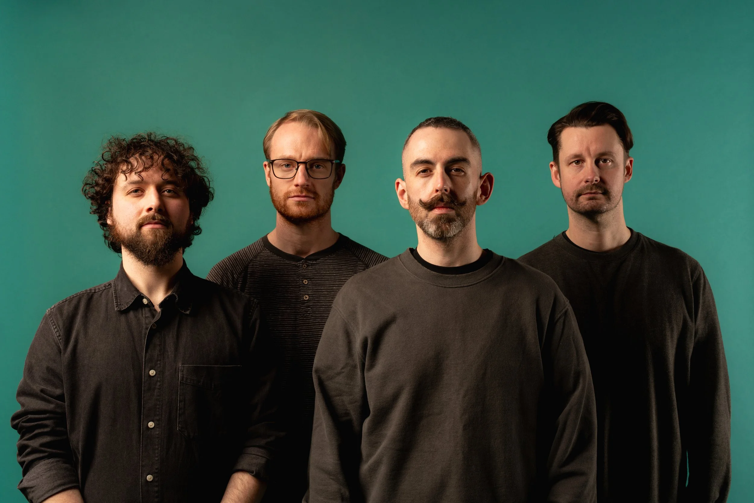 Four men standing side by side in front of a green background, wearing dark casual clothing, with serious expressions.