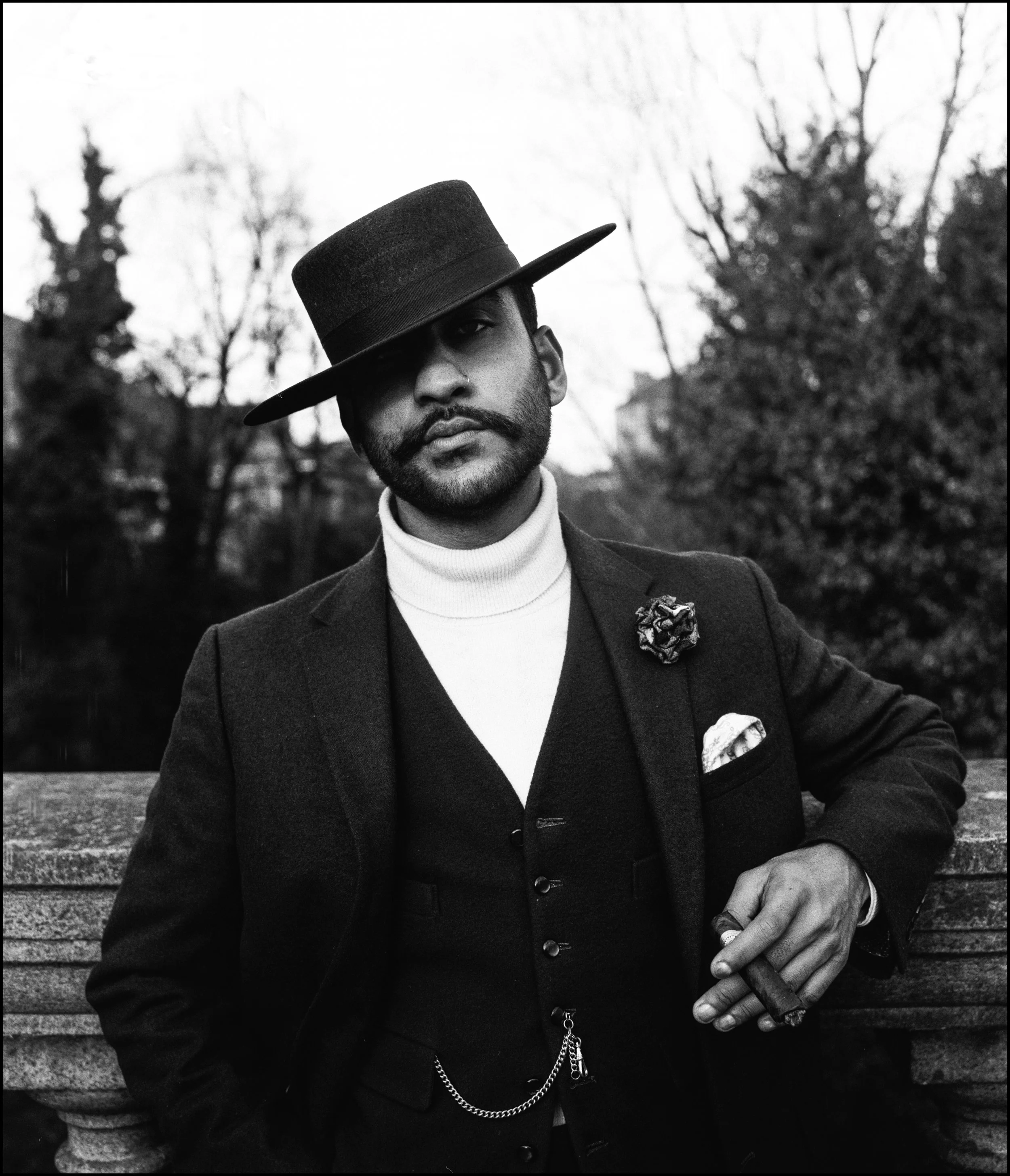 A man dressed in vintage style clothing, wearing a dark suit, a waistcoat, and a white turtleneck. He has a well-groomed beard, a mustache, and a dark hat tilted over one eye. He is holding a cigar in his right hand and stands in front of a park benc