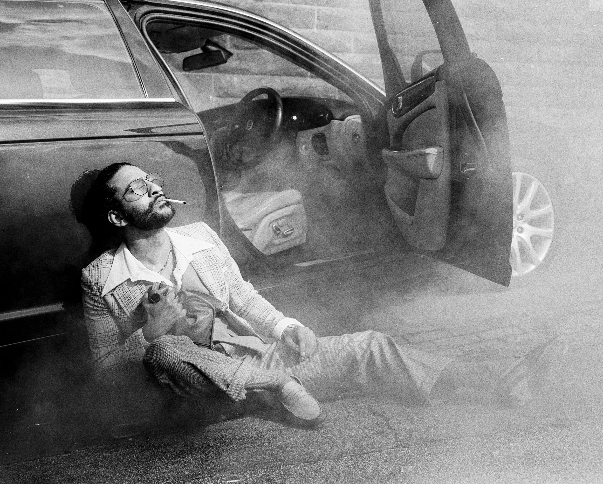 A man with glasses and a beard, wearing a plaid blazer, sits next to an open car door with smoke around him. He is smoking a cigarette and holding a bottle, with his legs stretched out on the ground.
