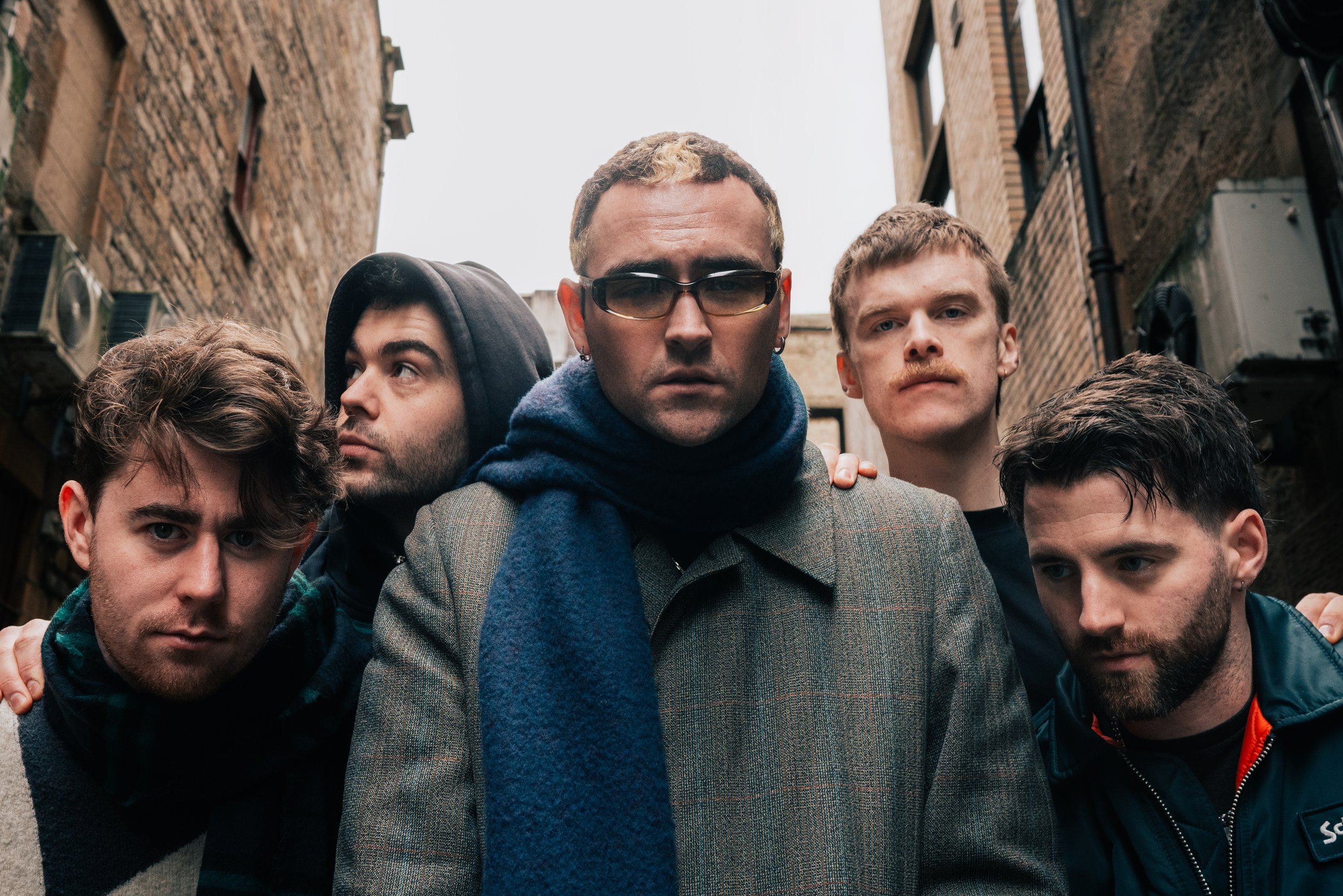 Group of five young men standing close together outdoors in an alleyway, with brick buildings on either side, looking serious and intense.