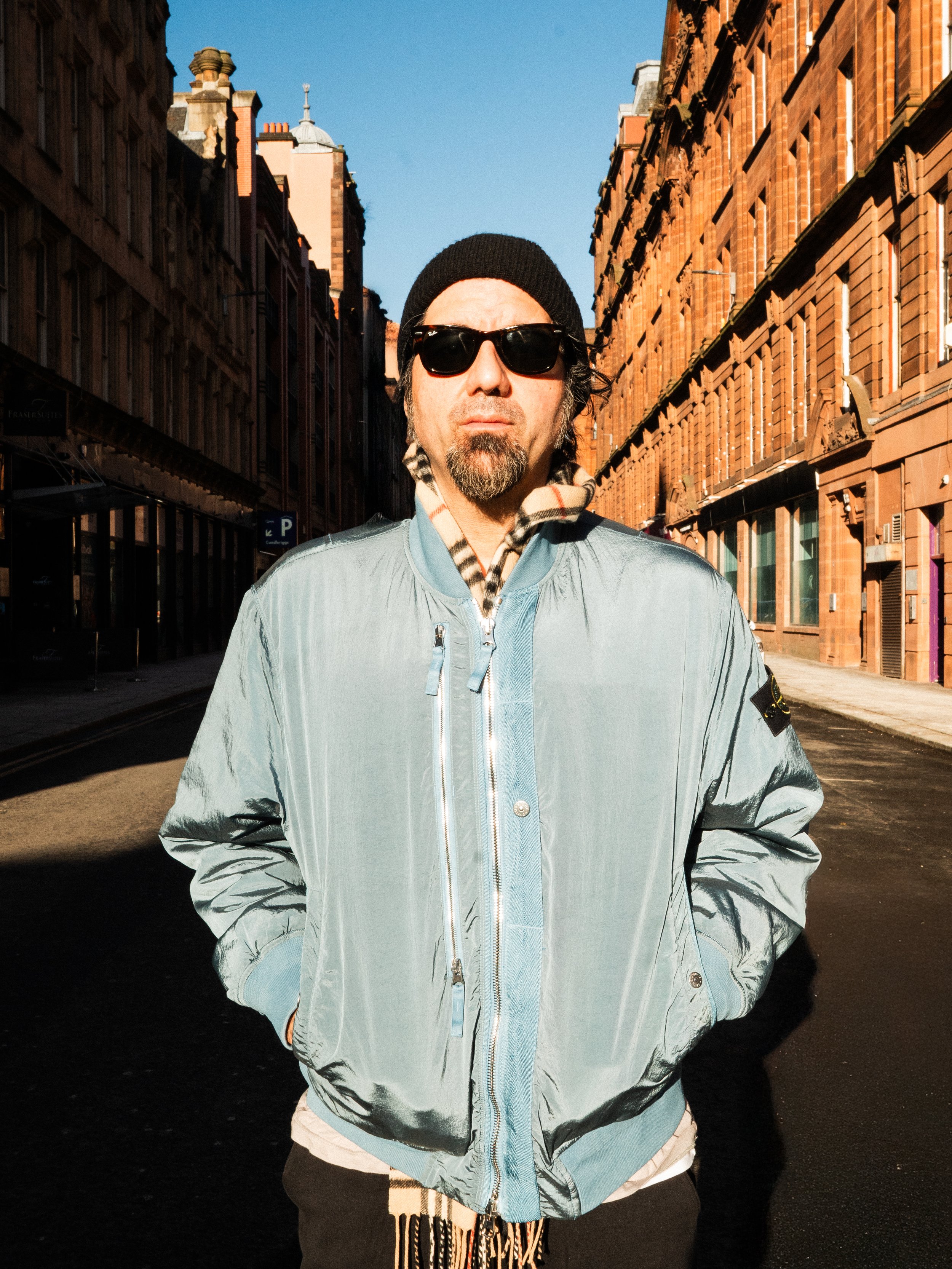 A man with a beard and goatee, wearing sunglasses, a beanie, a light blue jacket, and a patterned scarf, standing on a city street during daytime with old brick buildings on either side.