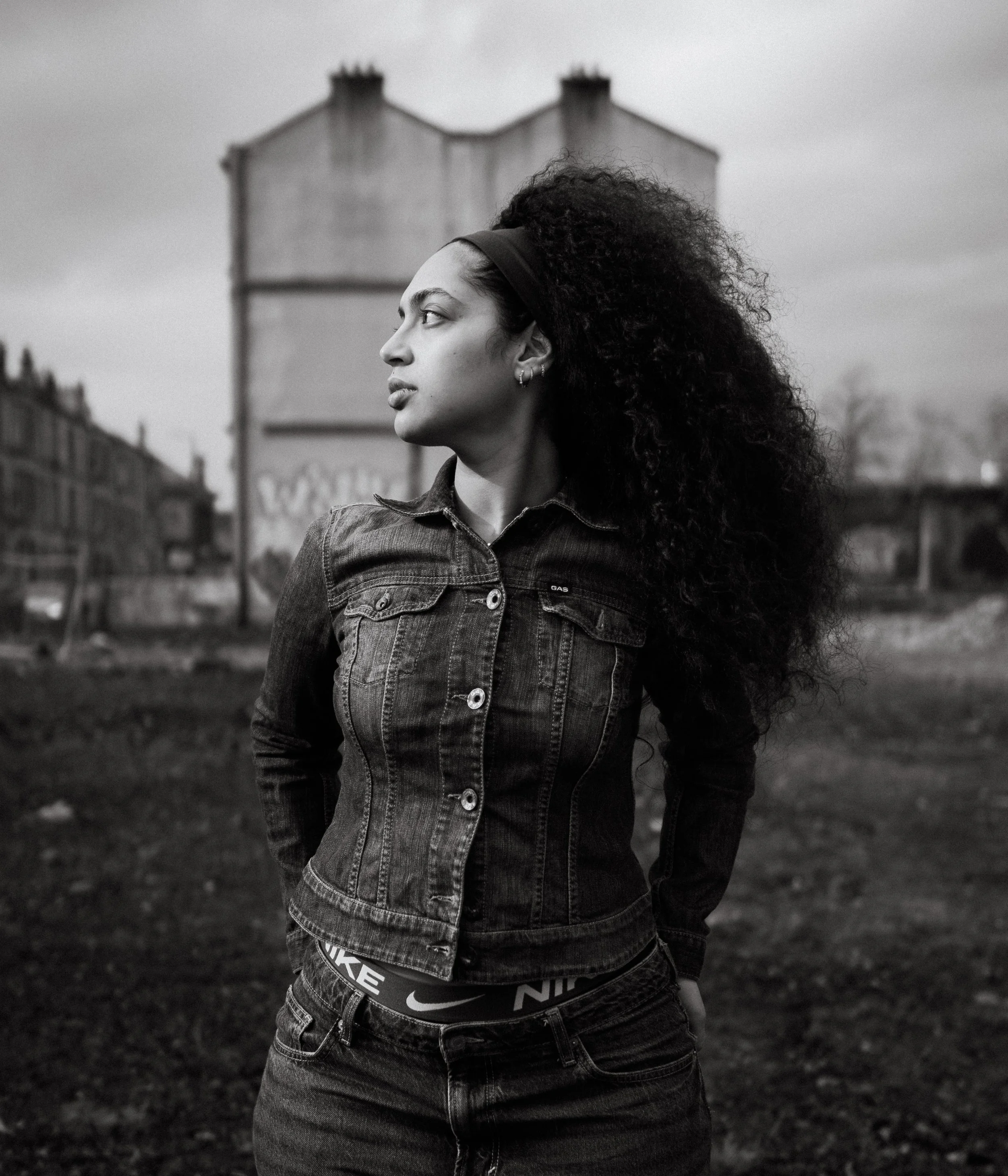 A young woman with curly hair and earrings stands outdoors in an urban setting, looking to the side, wearing a denim jacket and Nike underwear, with a background of buildings and a cloudy sky.
