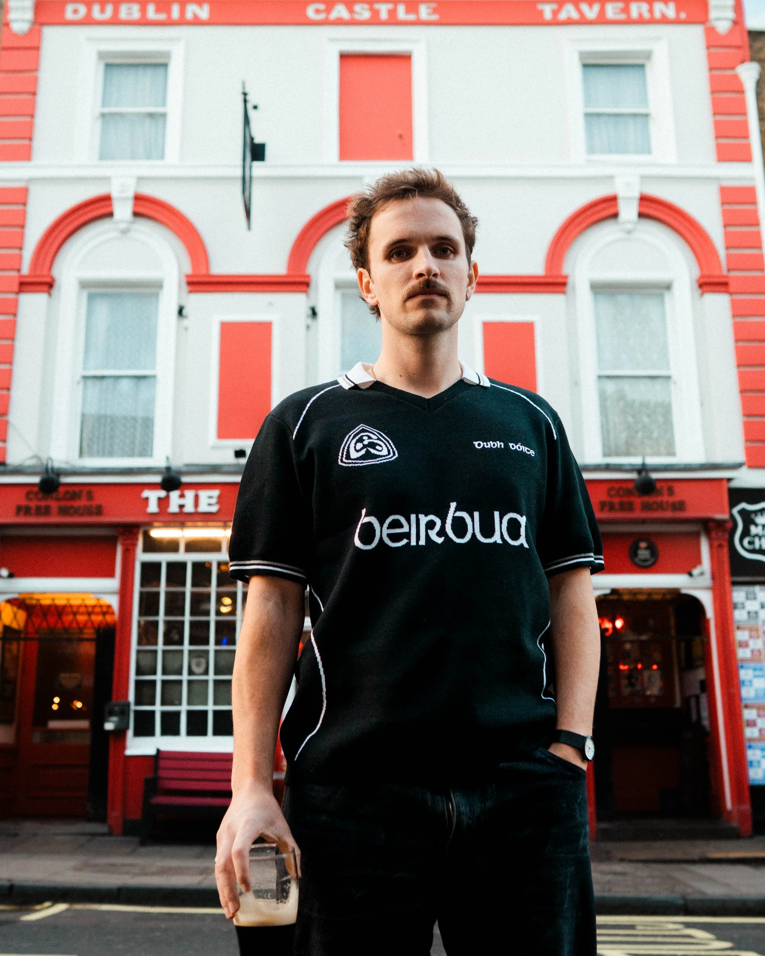 A young man stands in front of a red and white building with signs reading 'Dublin Castle Tavern' and 'The Cooden's Free House.' He wears a black sports jersey with white accents, holding a glass of dark beer, and looks directly at the camera.