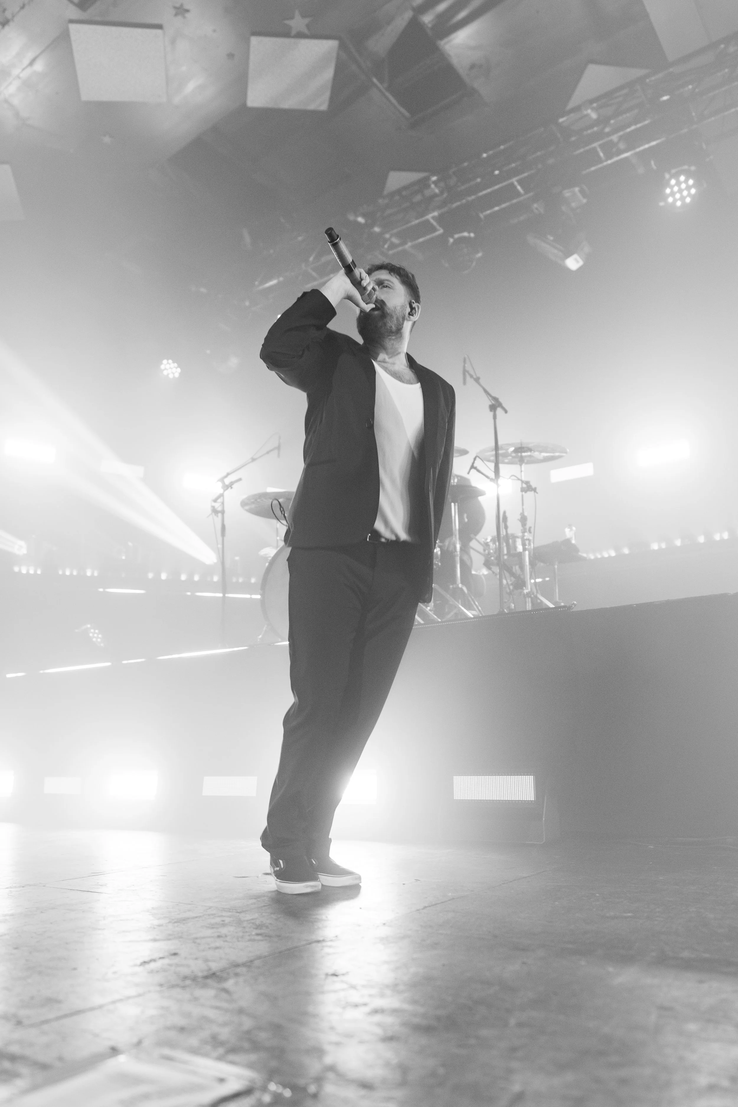 A male singer performs on stage, holding a microphone to his mouth, with a drum kit and stage lights in the background.