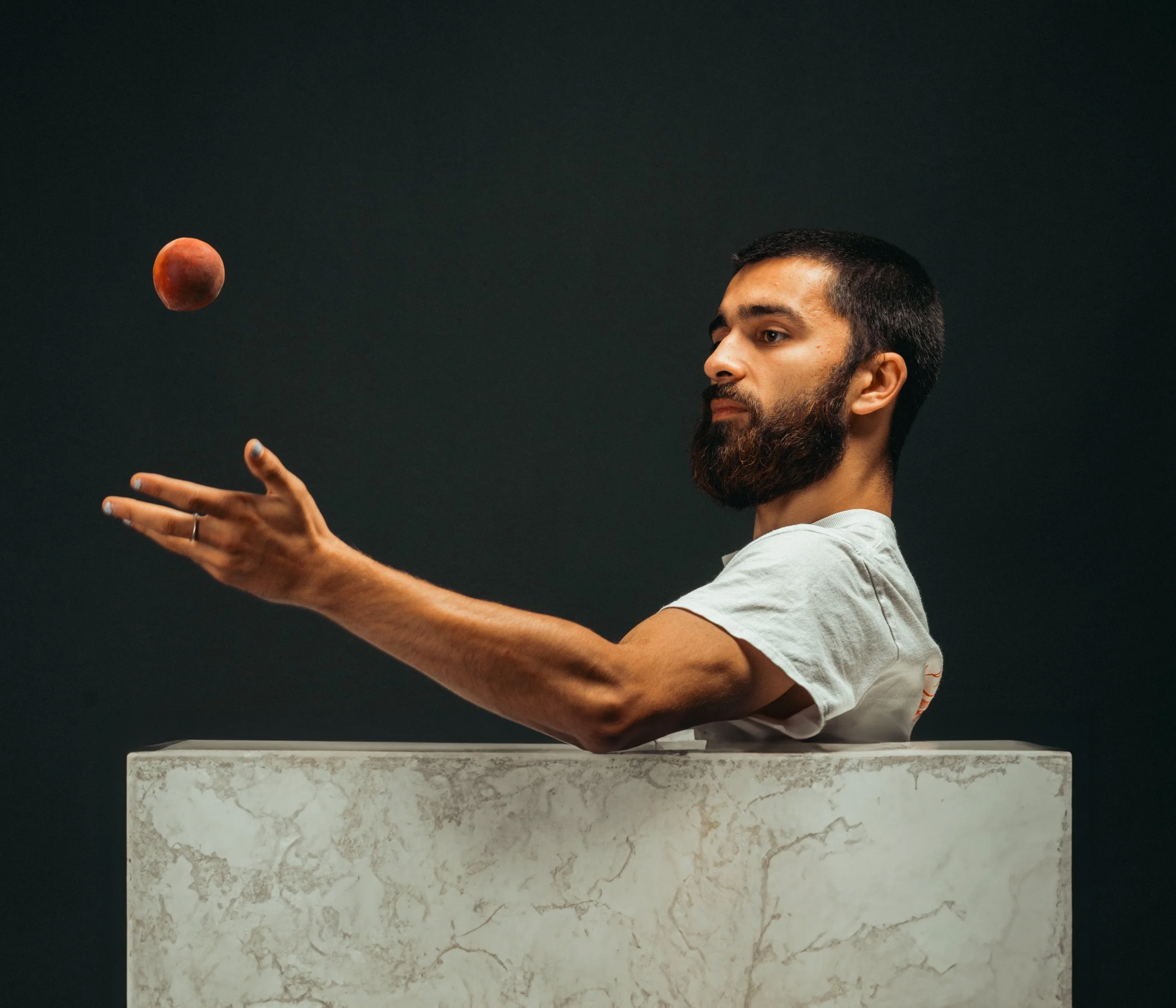 A man with a beard is sitting behind a white marble box, balancing a small, burnt orange moon-like object on his outstretched hand against a dark background.