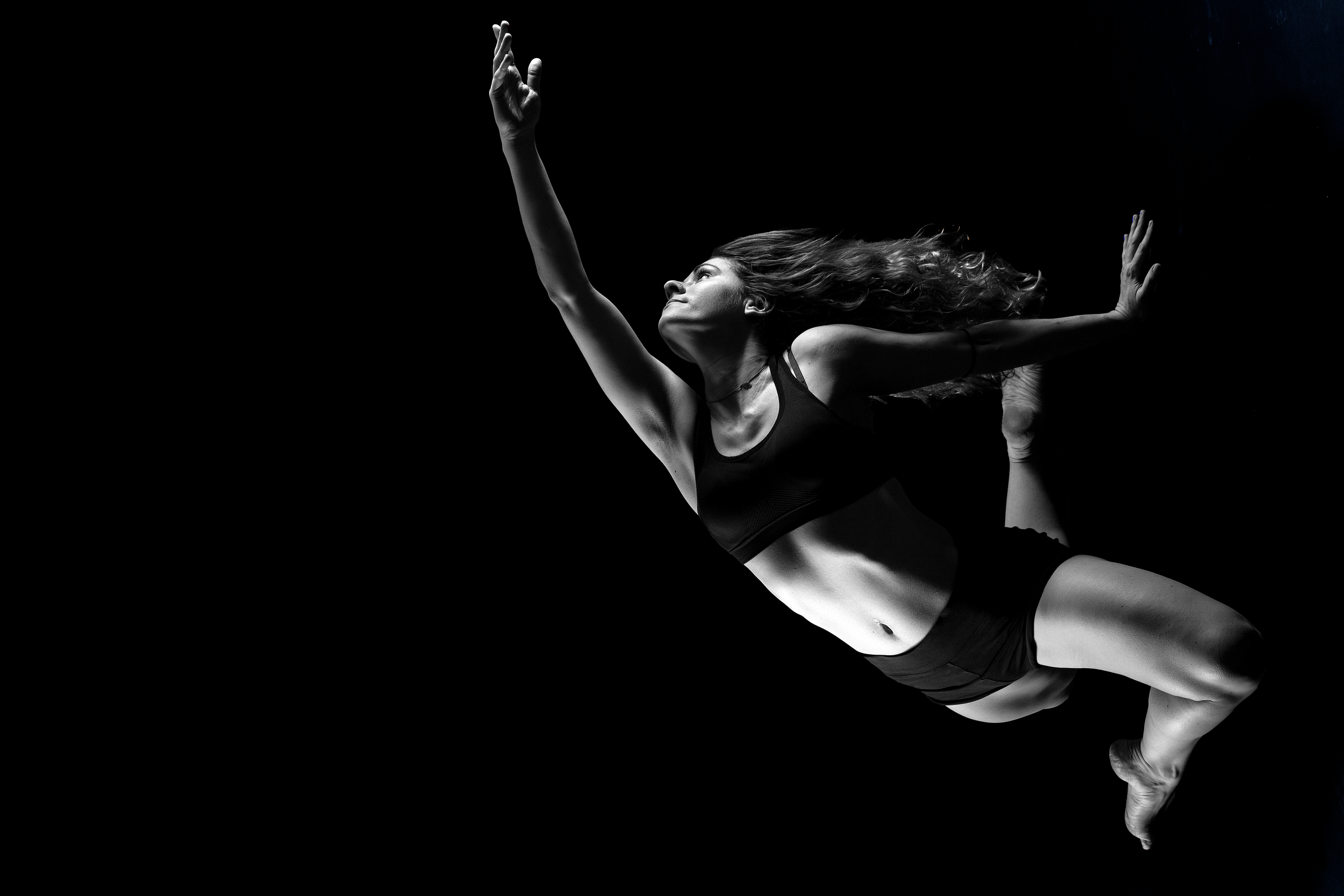 A woman performing a mid-air yoga pose against a dark background, captured in black and white.