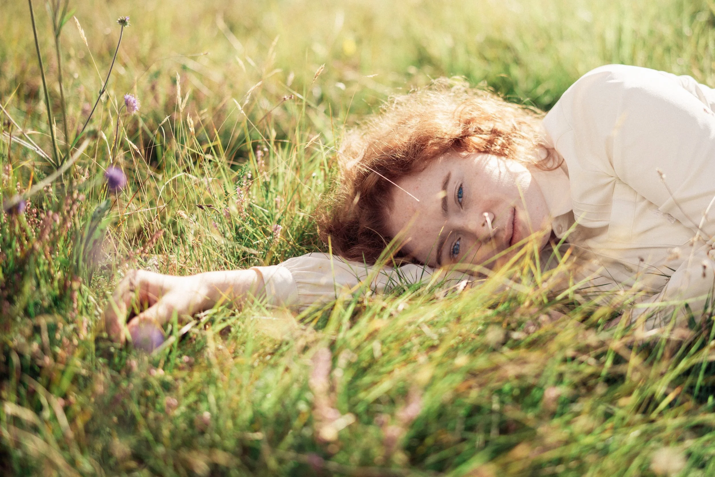 A woman with red hair and blue eyes wearing a white shirt lying in a grassy field with wildflowers, looking at the camera.