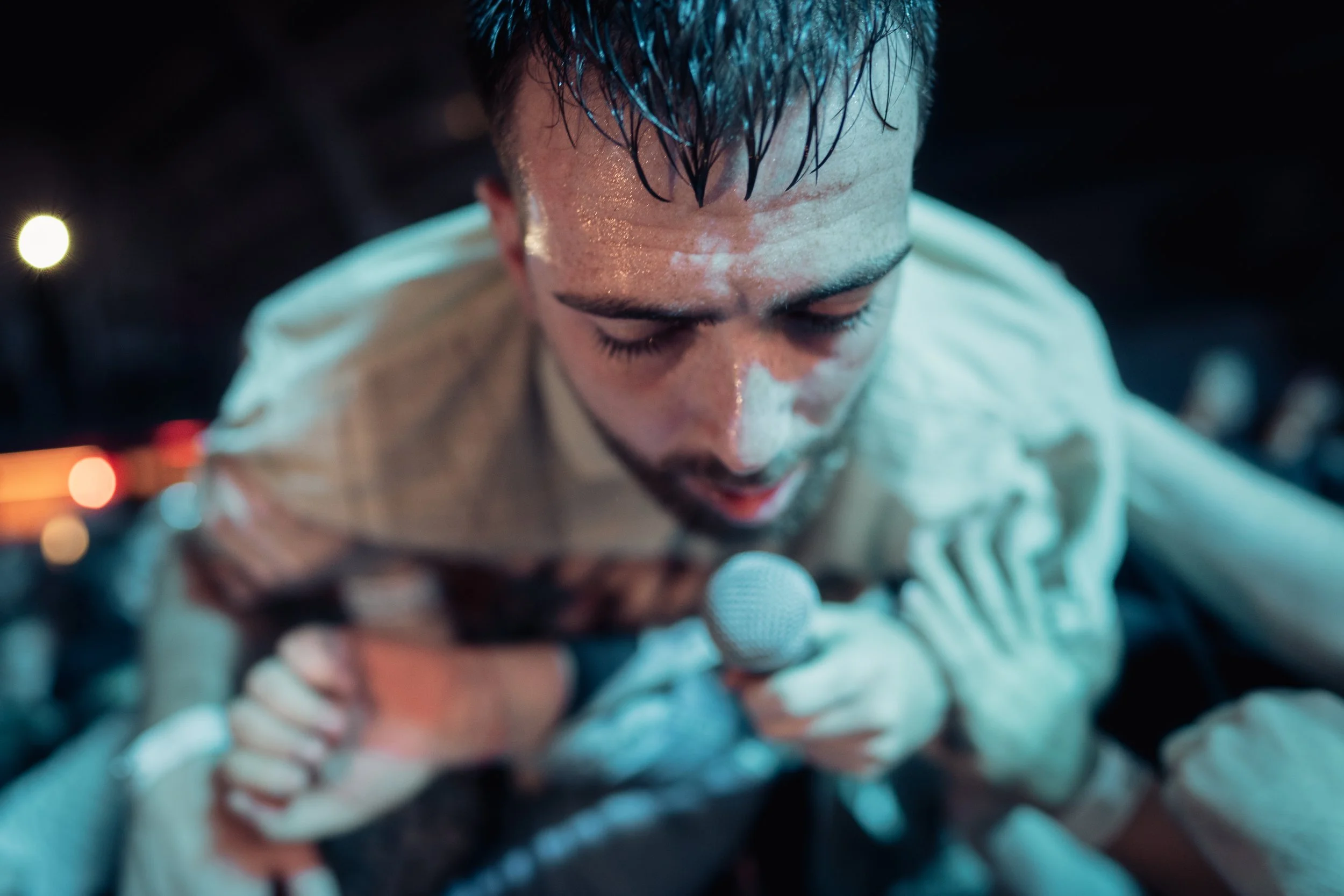 A man with wet hair and a beard, holding a microphone close to his mouth, appears to be singing or speaking passionately, with a dark background and blurred lights.