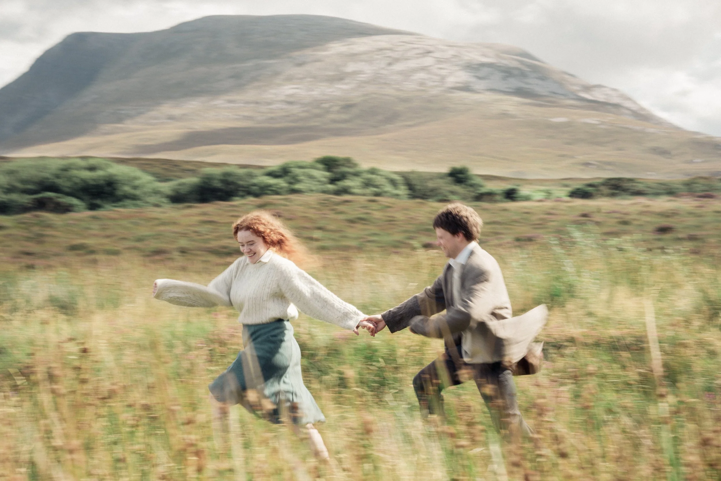 A young woman and a young man holding hands and running through a grassy field with mountains in the background, smiling and enjoying themselves.