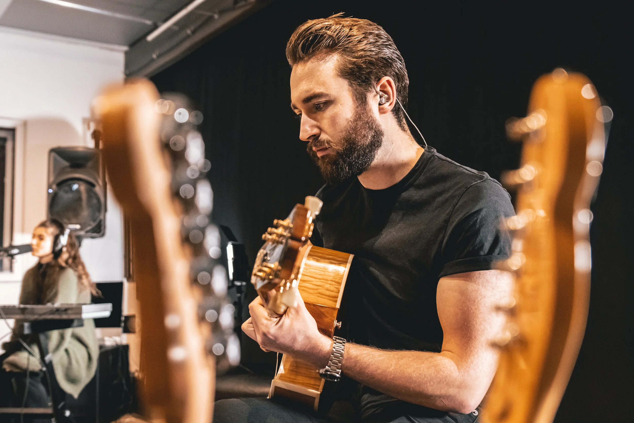 A bearded man in a black t-shirt playing an acoustic guitar during a music session, with a woman in the background sitting at a keyboard, in a dimly lit room.