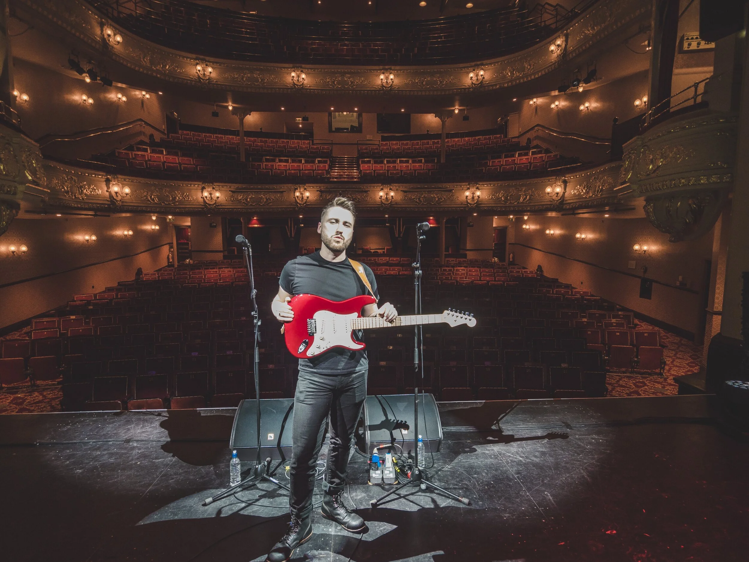 A male musician holding a red electric guitar standing on stage in an empty theater or auditorium with ornate balconies and warm lighting.