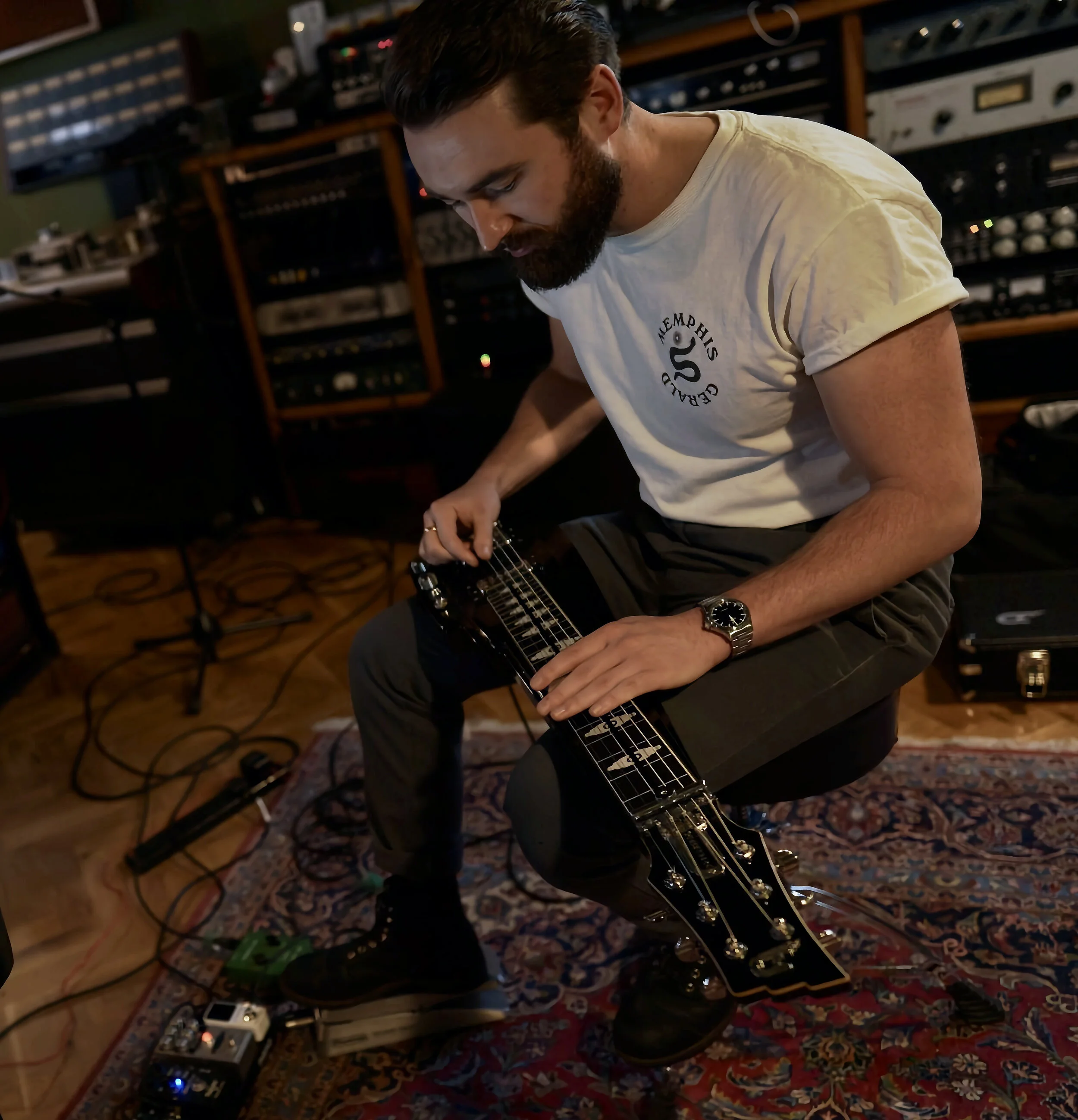 Man with a beard playing a lap steel guitar in a recording studio, wearing a white t-shirt and black pants, sitting on a stool.