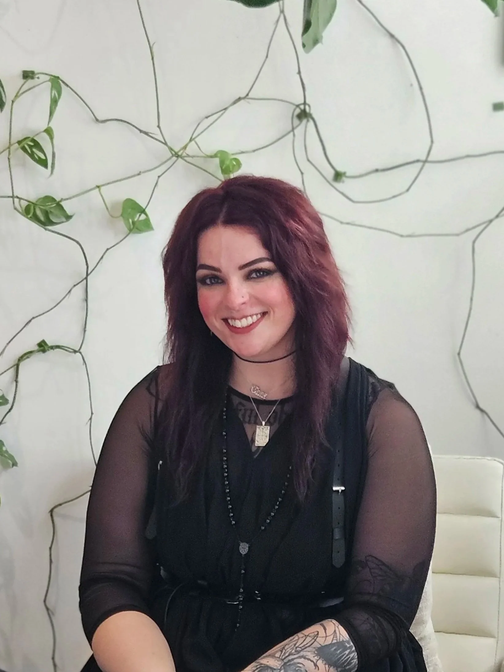 A woman with dark hair styled in long braids, wearing a black outfit with a large bow tie, standing in front of a green plant with large leaves.