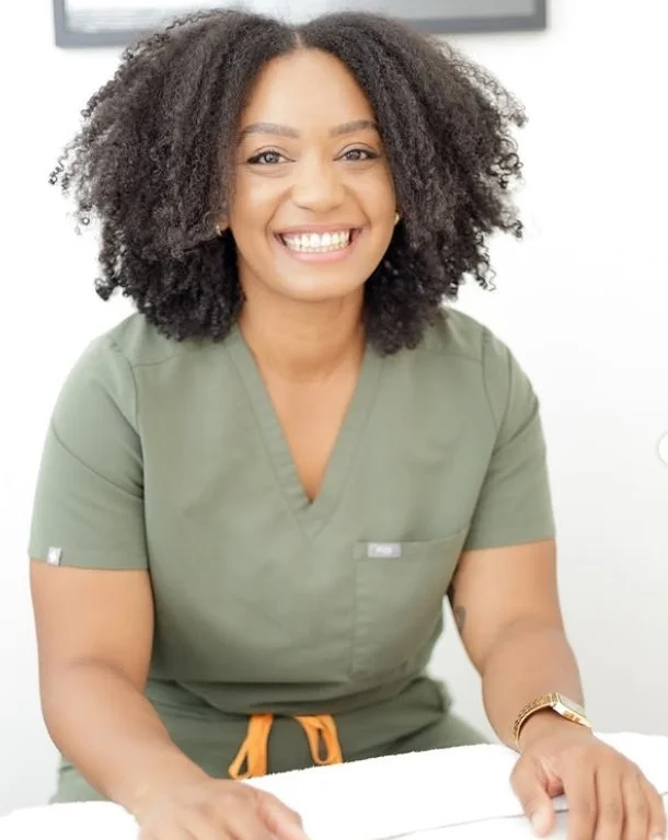 A woman with curly hair, glasses, wearing black, is giving a facial massage to a person lying down with closed eyes in a beauty or skincare treatment room.