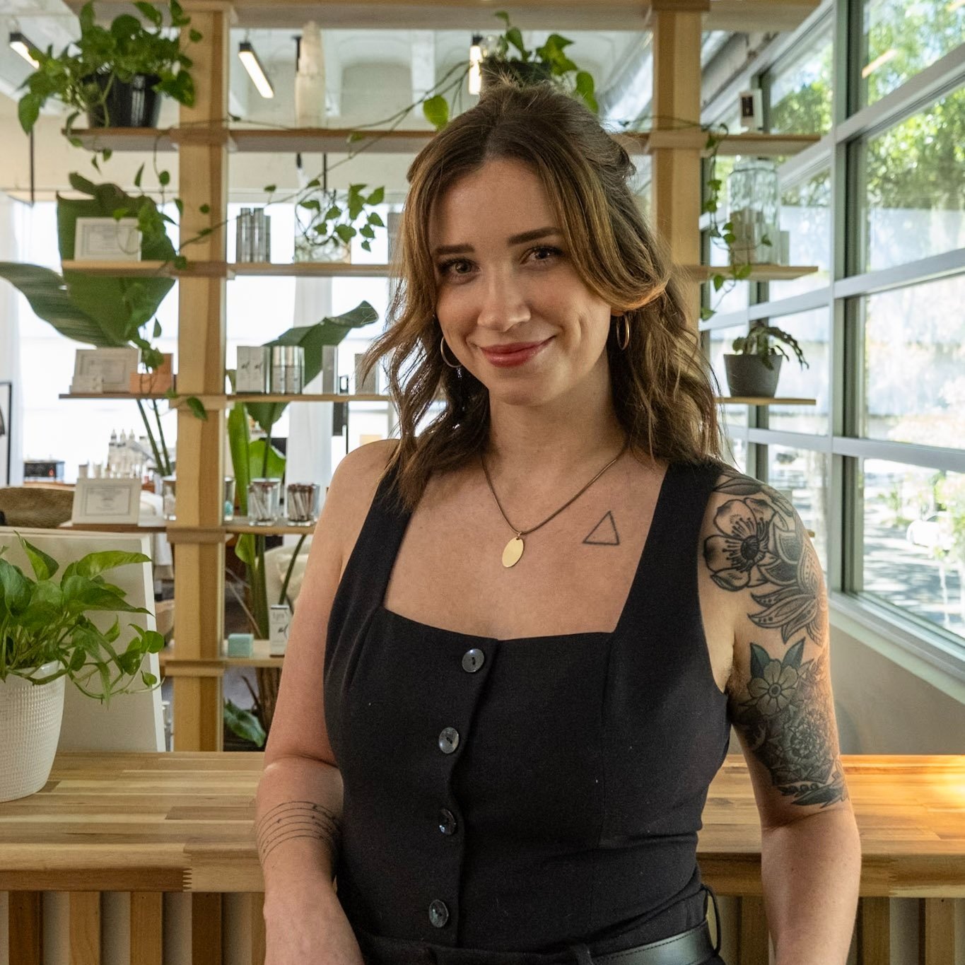 A woman with shoulder-length brown hair, wearing a black sleeveless top with buttons, standing indoors near a wooden counter. She has tattoos on her right shoulder and arm, wears a gold necklace with a round pendant, and has hoop earrings. She is smiling and looking at the camera, with large windows and indoor plants in the background.