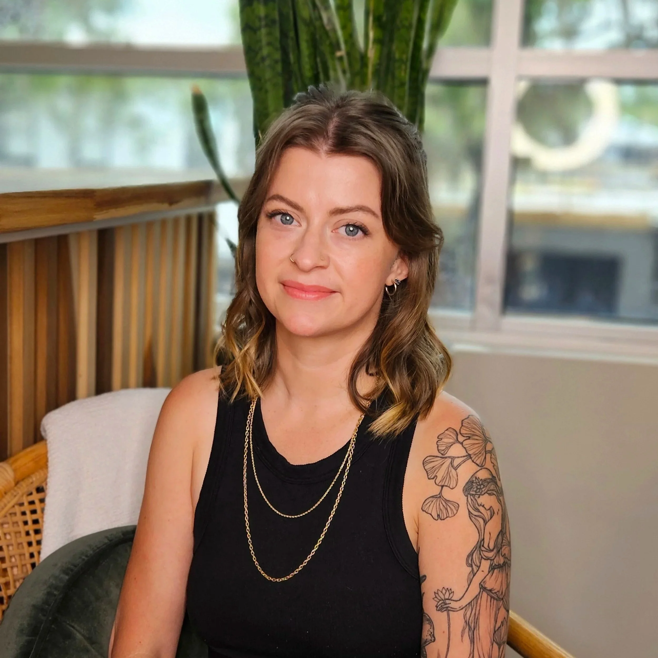 A young woman with shoulder-length wavy brown hair, blue eyes, and light skin, wearing a black sleeveless top, layered gold necklaces, and earrings, sitting indoors near a large green plant and a window with a view.