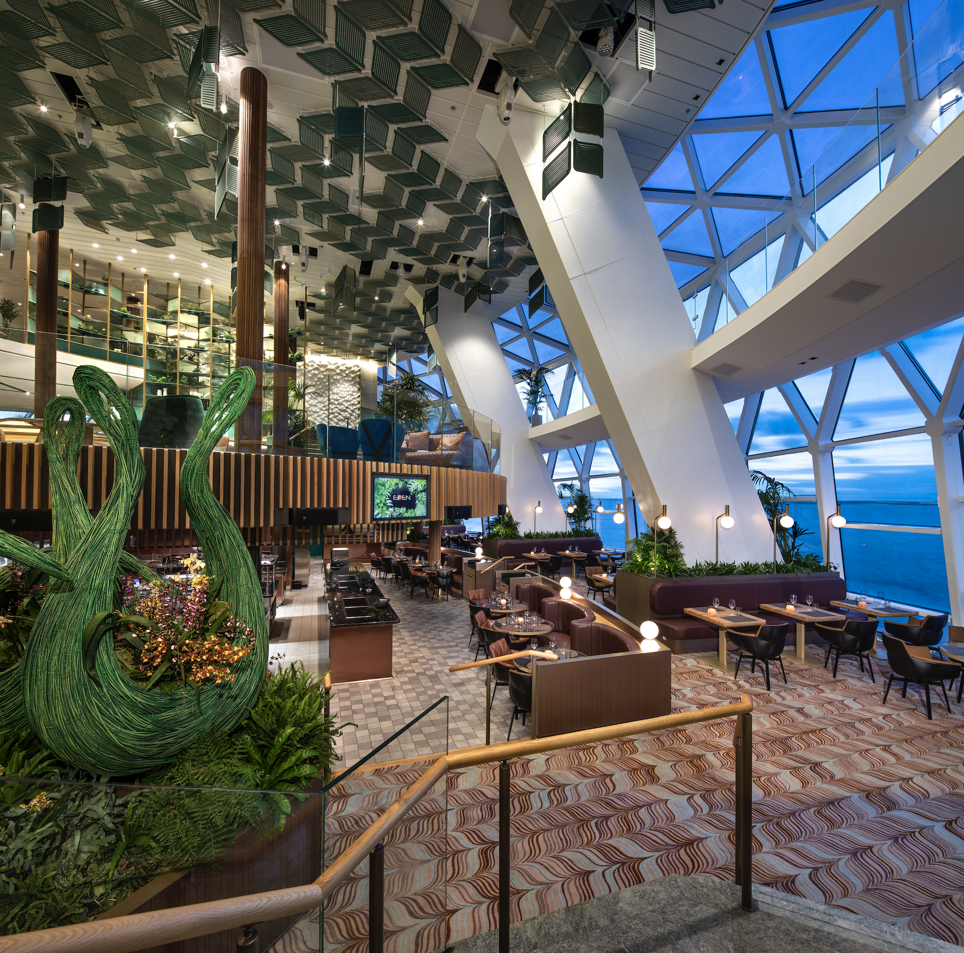 Interior of a modern restaurant with large windows offering an ocean view, featuring contemporary furnishings, green and wooden decor elements, a unique ceiling design, and ambient lighting. Tables are set for dining with some areas decorated with pl