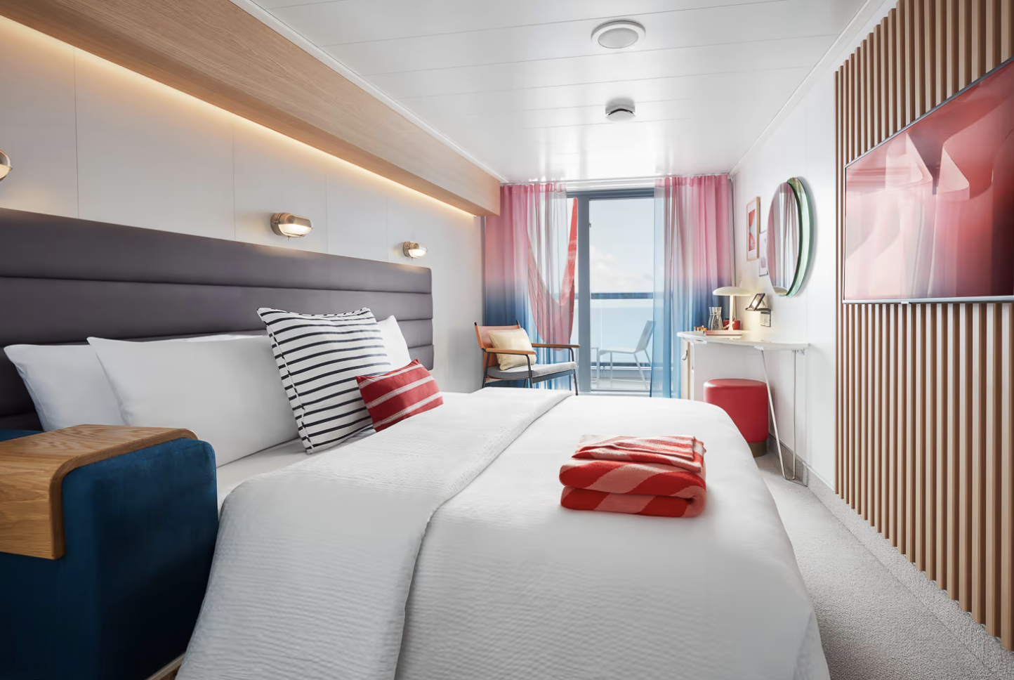 Modern hotel room with a large bed, striped pillows, and red accents, featuring a balcony with ocean view, a desk, chair, and a wall-mounted TV.