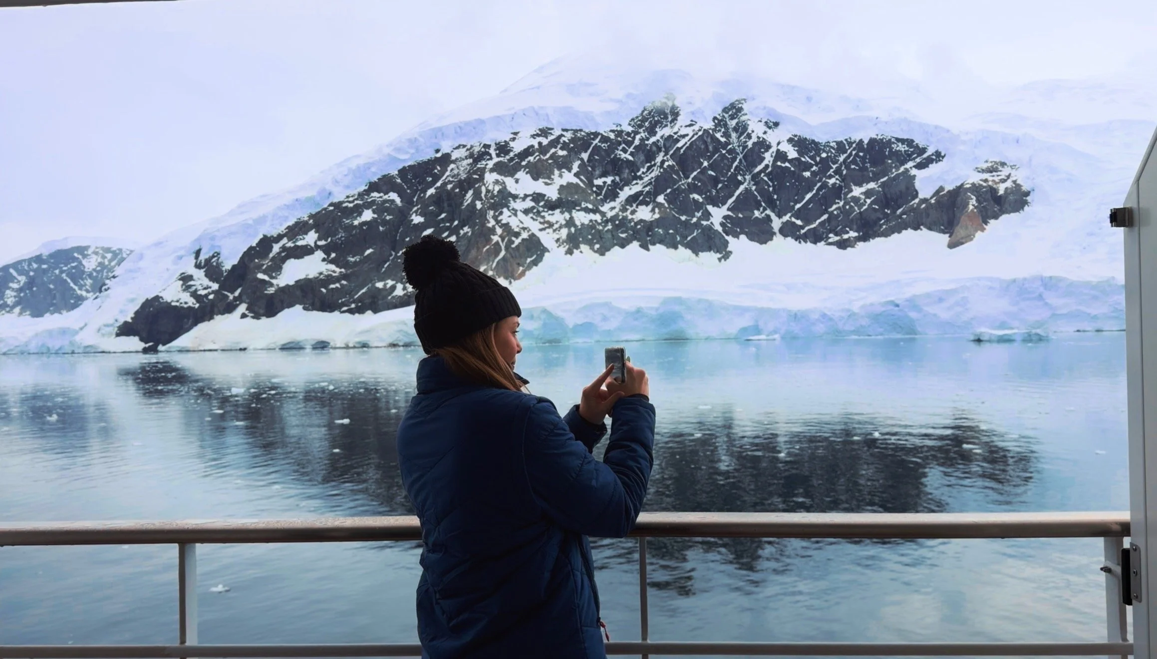 Woman standing on balcony of expedition ship in Antarctica taking a video of the stunning snow covered glaciers.
