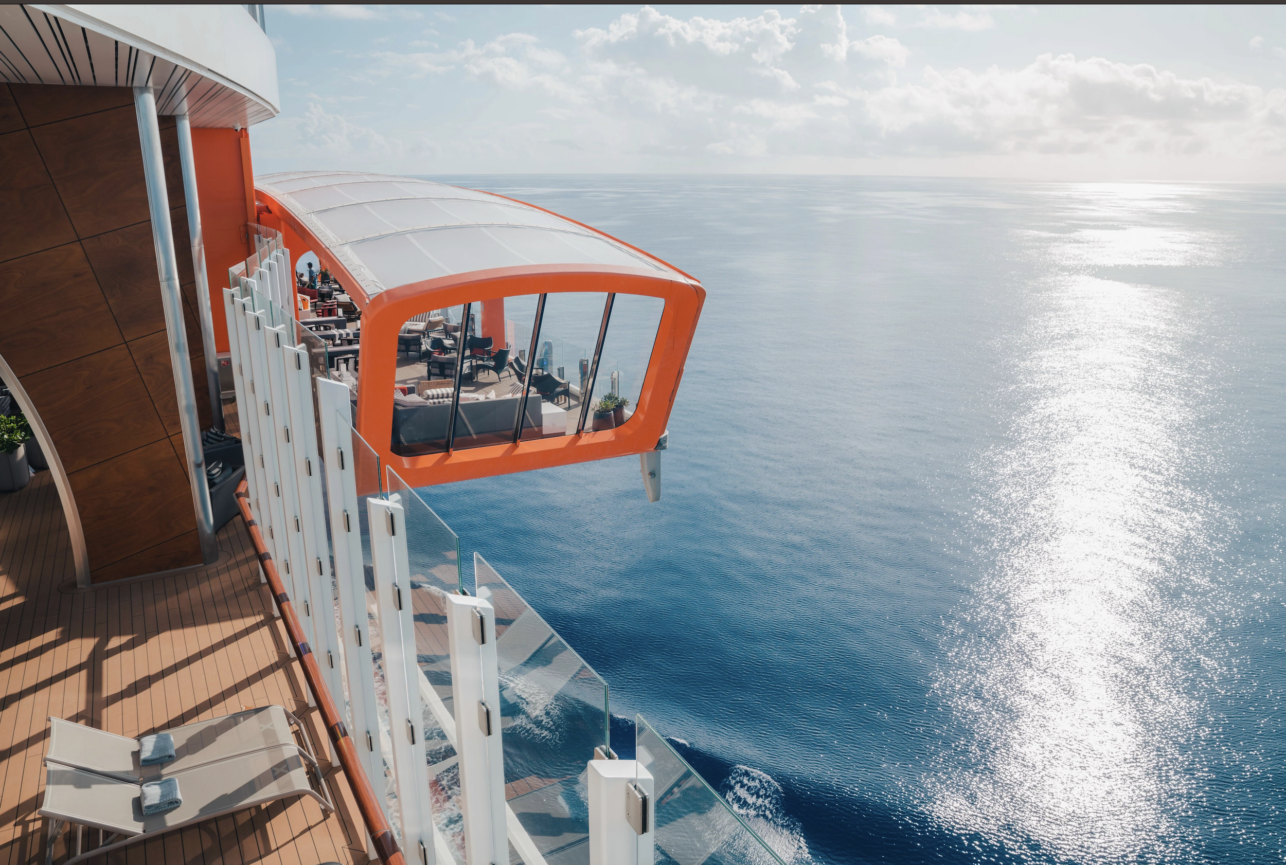 Aboard a cruise ship with a modern, orange, semi-enclosed lounge extending over the ocean; clear blue sky and calm sea visible.