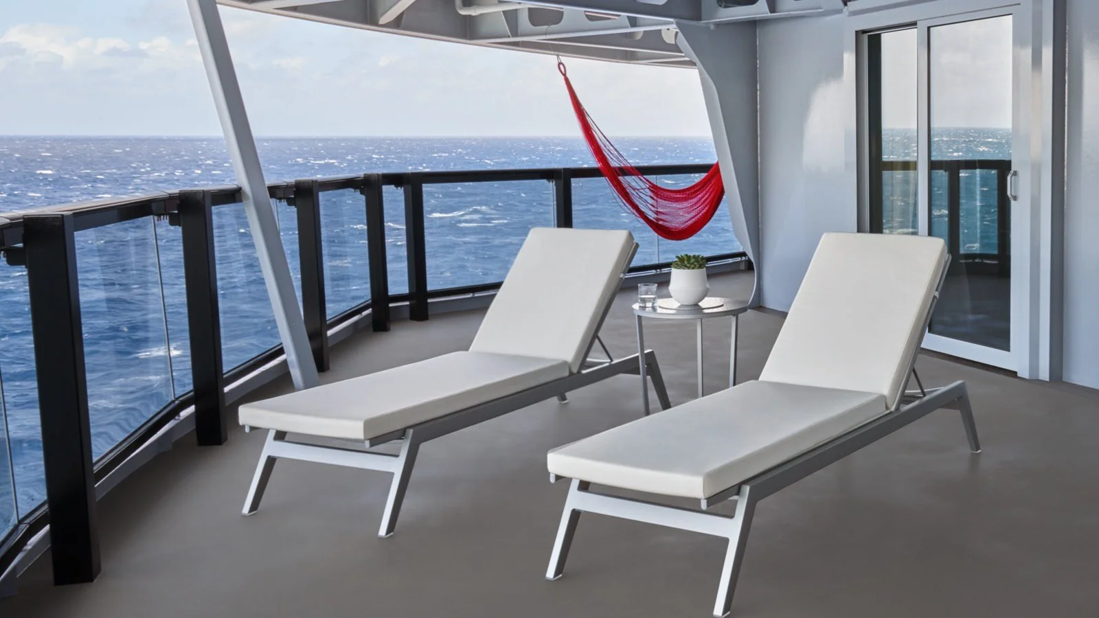 Cruise ship balcony with two lounge chairs, a hammock, small table, and ocean view.