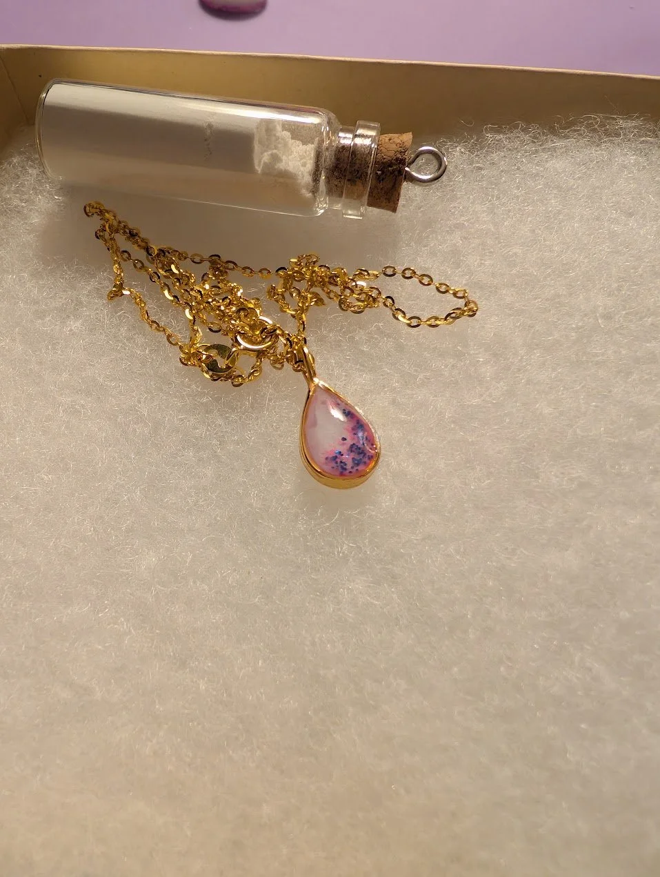 A gold necklace with a teardrop-shaped pink and purple pendant, placed on a soft white surface, with a glass vial and cork stopper nearby.