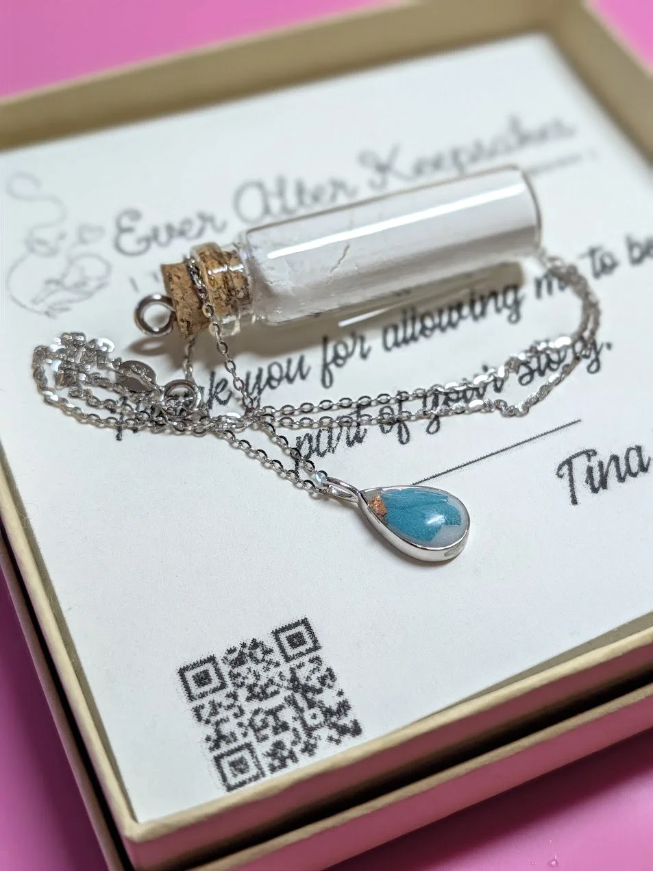 A necklace with a blue teardrop pendant and a small glass vial charm, placed on a white card inside a pink box.
