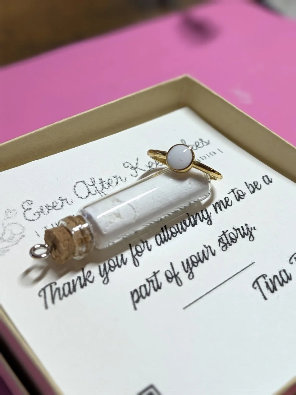 A ring with a white circular stone resting on a thank you note in a pink box. The note expresses gratitude for allowing the recipient to be part of a story.
