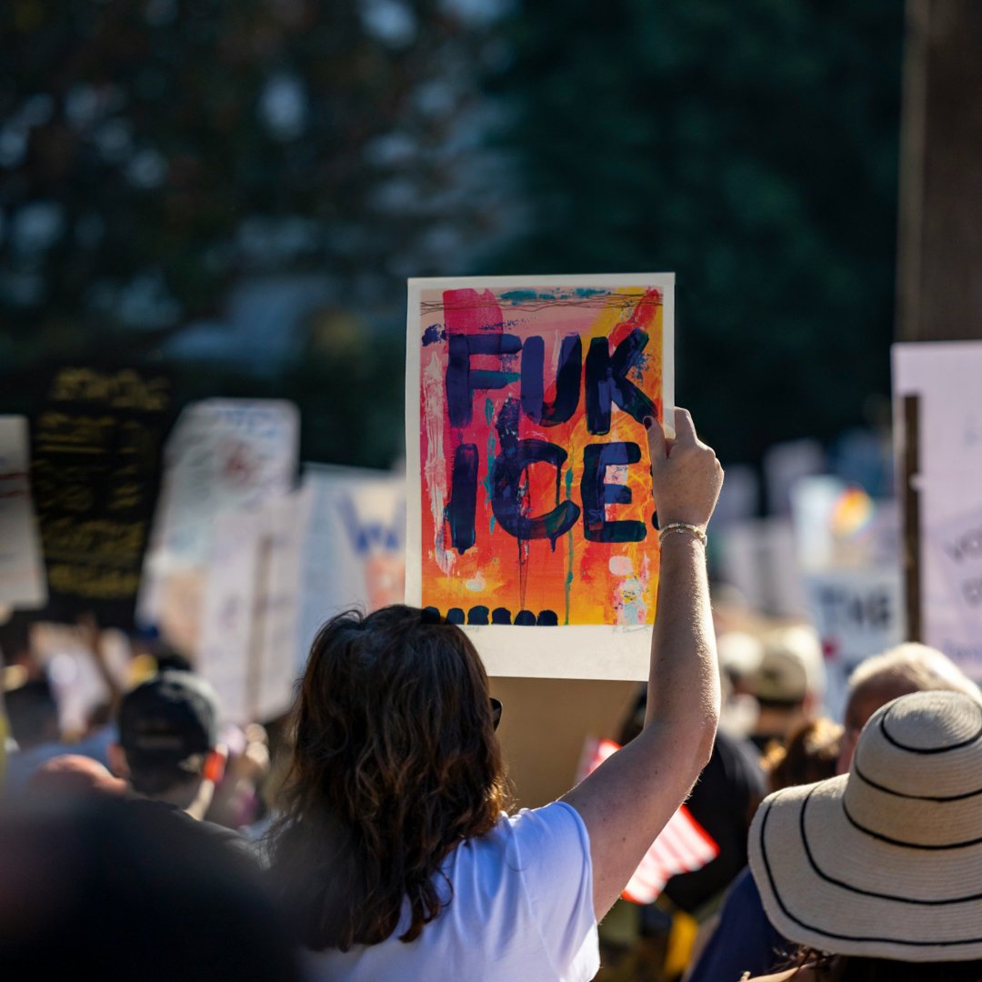 Person holding an 11×17 FUK ICE protest sign outdoors in a demonstration setting.  Background image adapted from Unsplash by Leo_visions